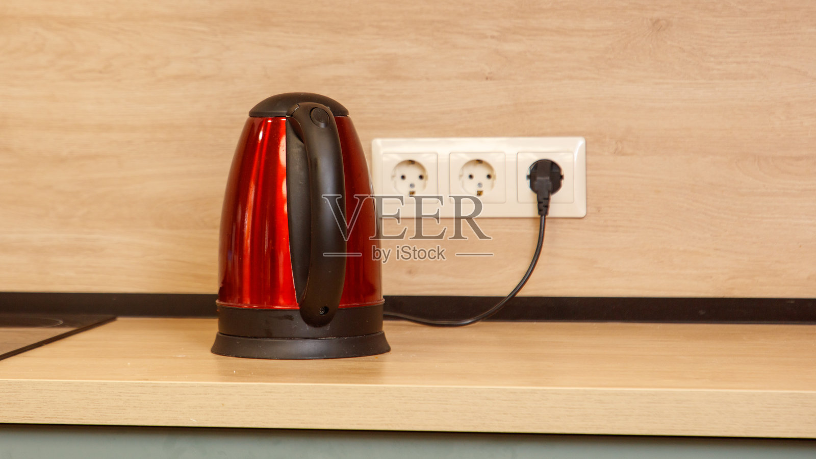 一个红色的电热水壶放在木制台面上,旁边有一个电源插座照片摄影图片