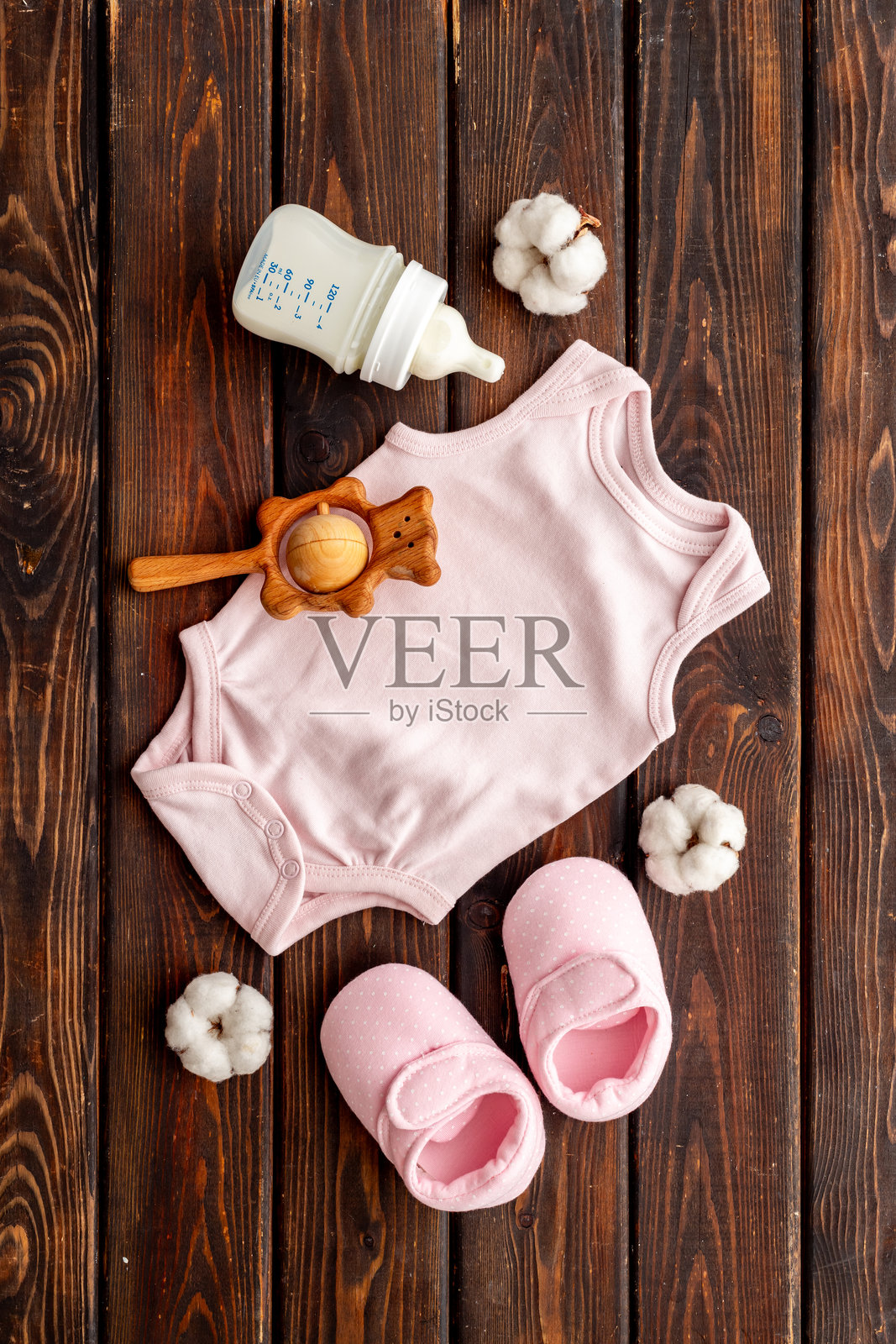 俯视角度拍摄的婴儿用品平铺照:婴儿连体衣、鞋子、木制牙胶玩具和奶瓶,背景为木质桌面。照片摄影图片