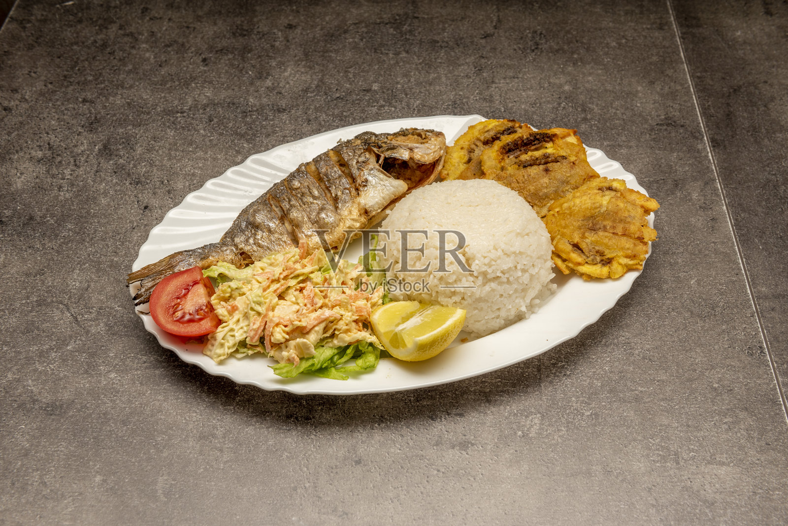 带有金头鲷鱼炸鱼、凉拌卷心菜、炸芭蕉片、白米饭和柠檬酱的精美烤盘,放在灰色桌子上照片摄影图片