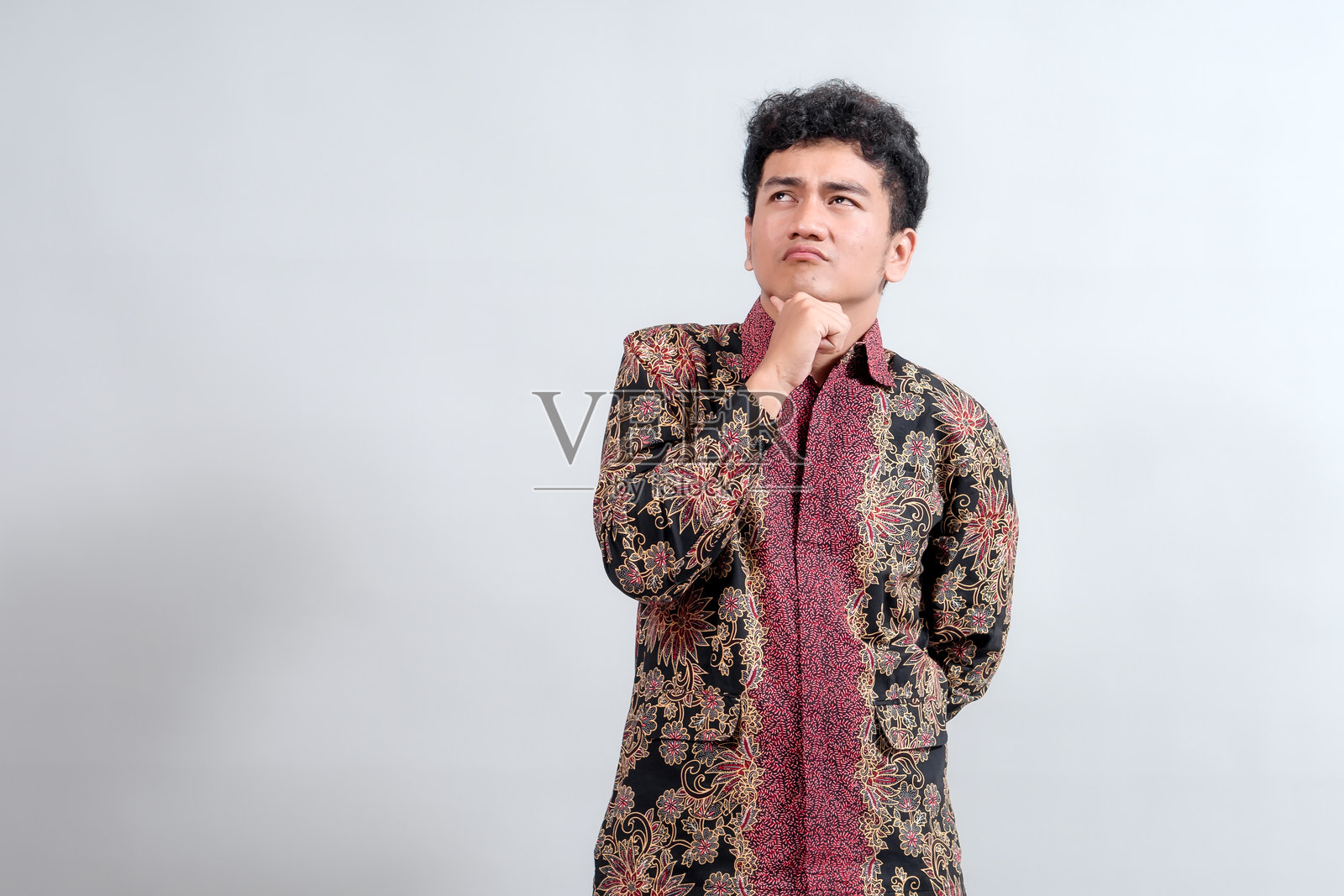 一位身穿传统蜡染衬衫的年轻男子,手托下巴,抬头沉思,非常适合文化或生活方式主题。照片摄影图片