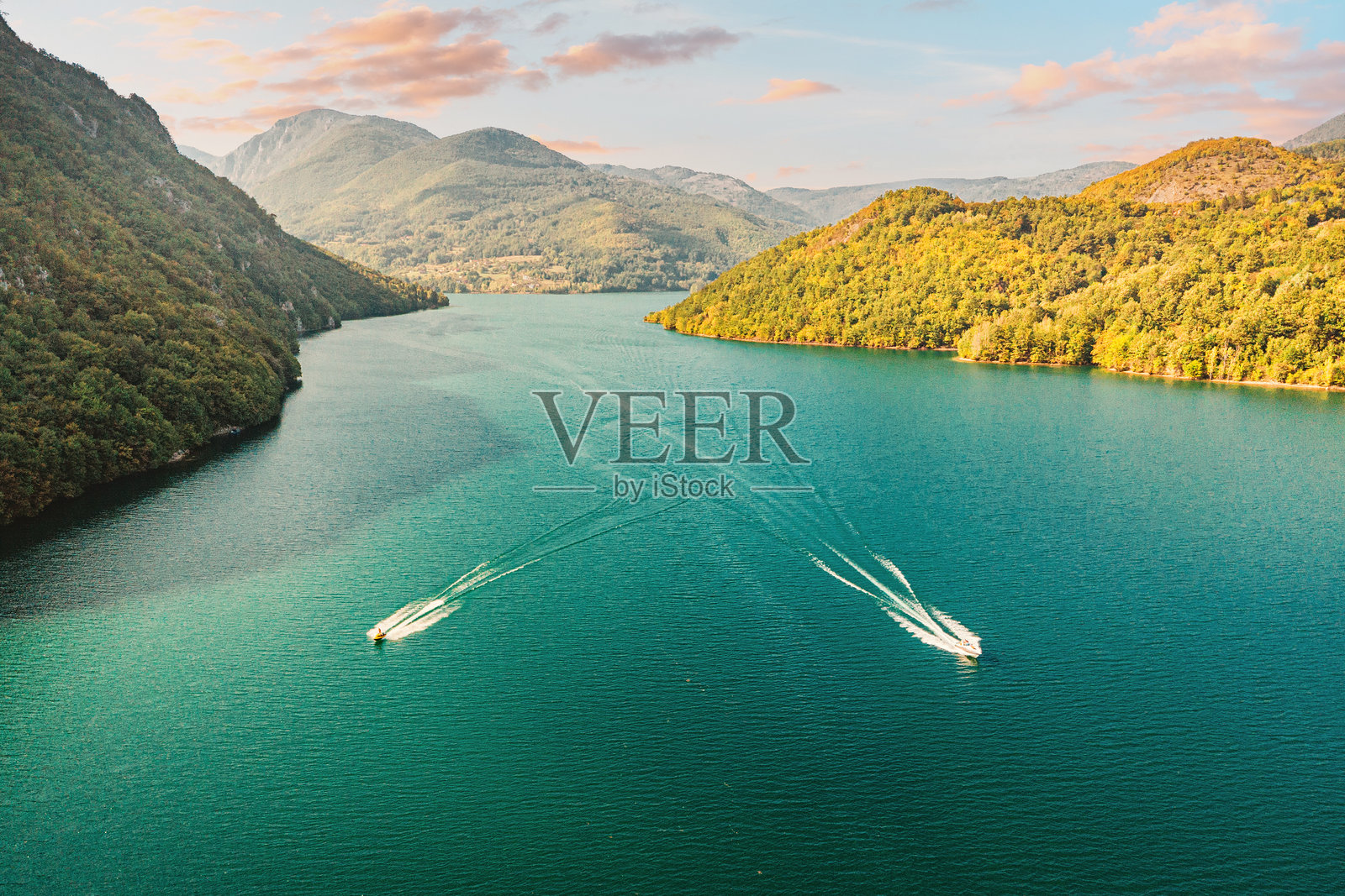 两艘快艇在一片广阔的绿松石色湖面上飞驰,身后留下了白色的航迹,四周环绕着森林覆盖的山峦,天空晴朗。照片摄影图片