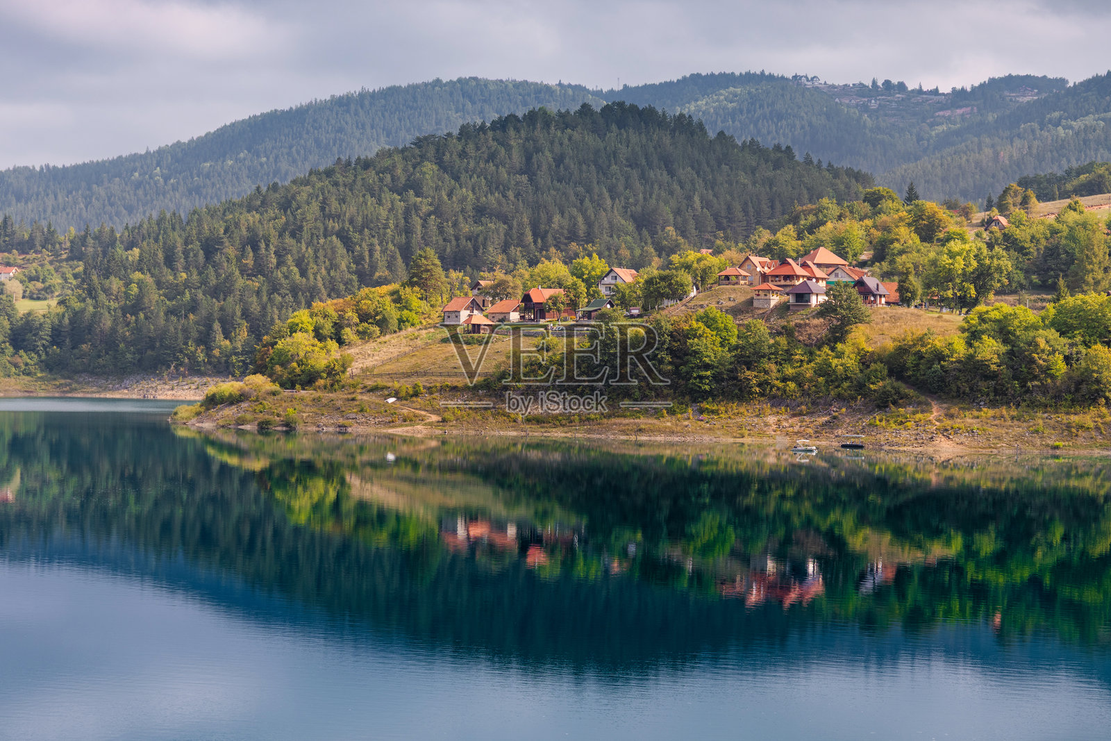 山坡上红顶房屋的村庄倒映在宁静的蓝色山湖水中,四周环绕着翠绿的森林。照片摄影图片