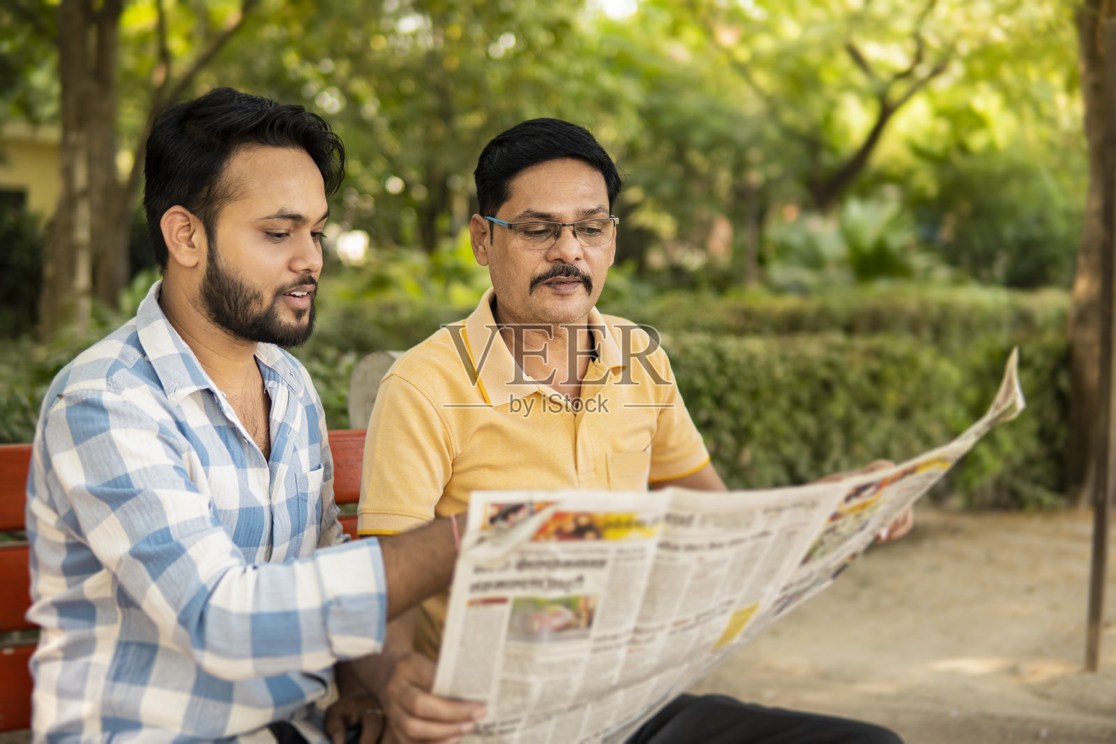 一位成熟的印度父亲和年幼的儿子在新鲜空气中,一同坐在公园长椅上阅读报纸。照片摄影图片