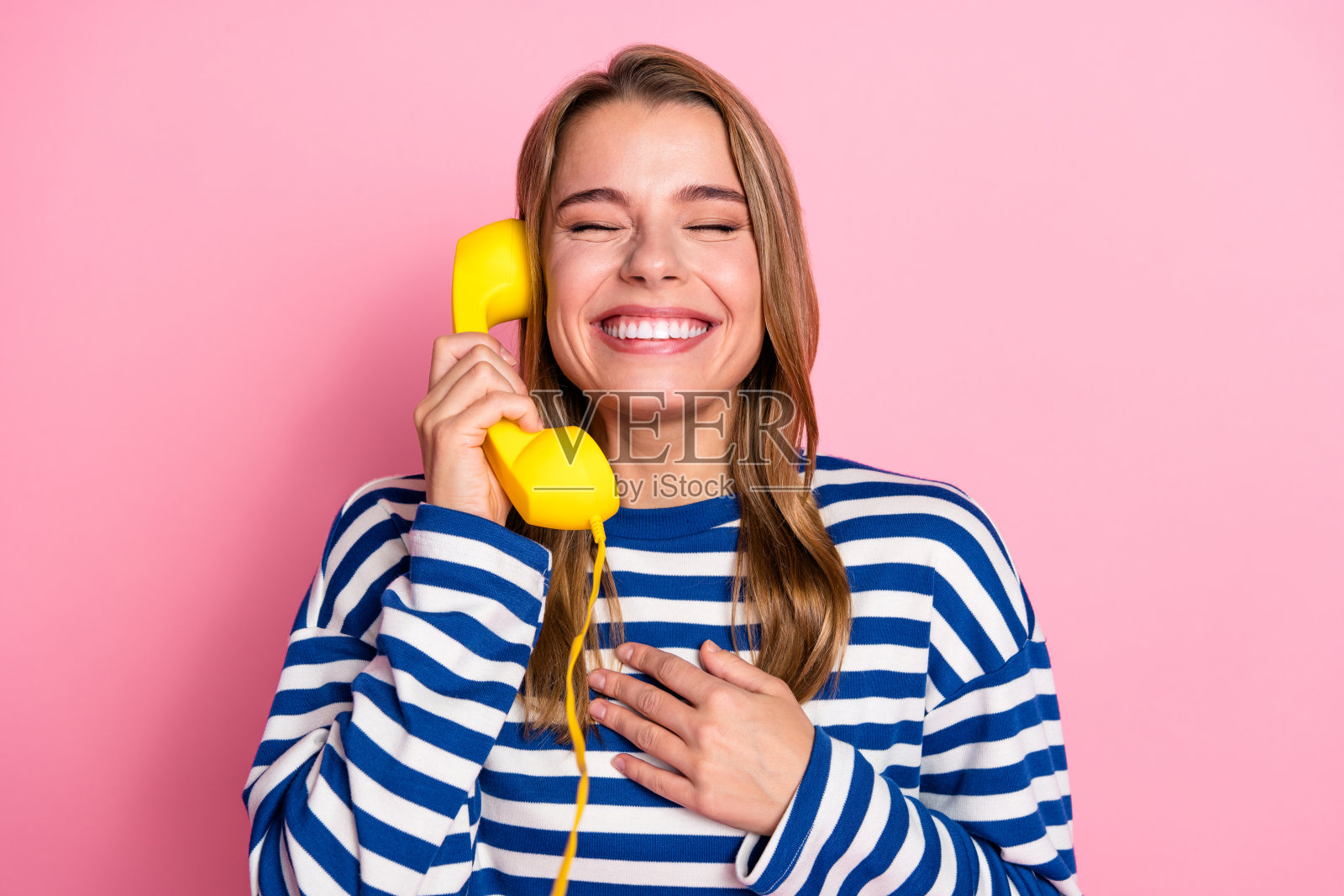 一位年轻快乐的女士,穿着休闲条纹毛衣,对着粉色背景微笑,手里拿着一部复古电话照片摄影图片