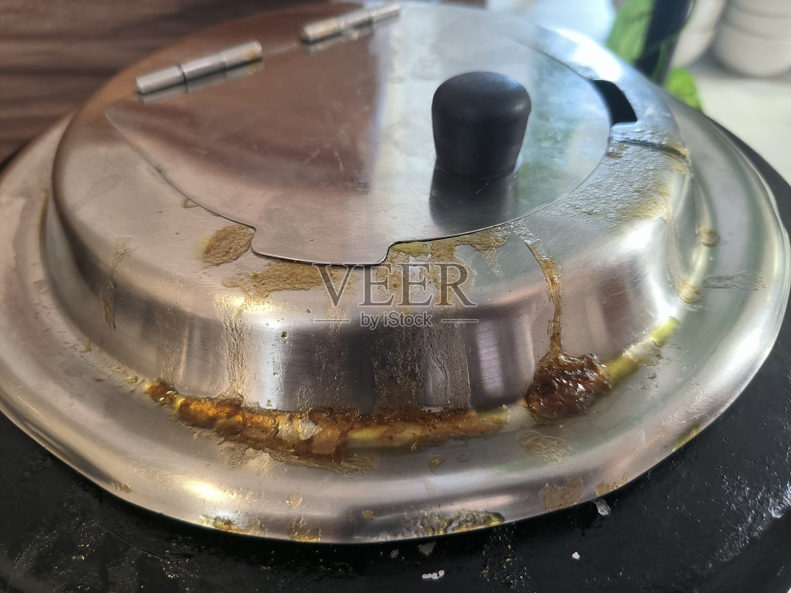 略微俯视的特写镜头,展示了一个黑色圆形餐盘或保温炉上的拉丝不锈钢盖子。盖子上有明显深棕色和黄色的烹饪残留物或酱汁污渍,尤其是在边缘和把手区域。照片摄影图片