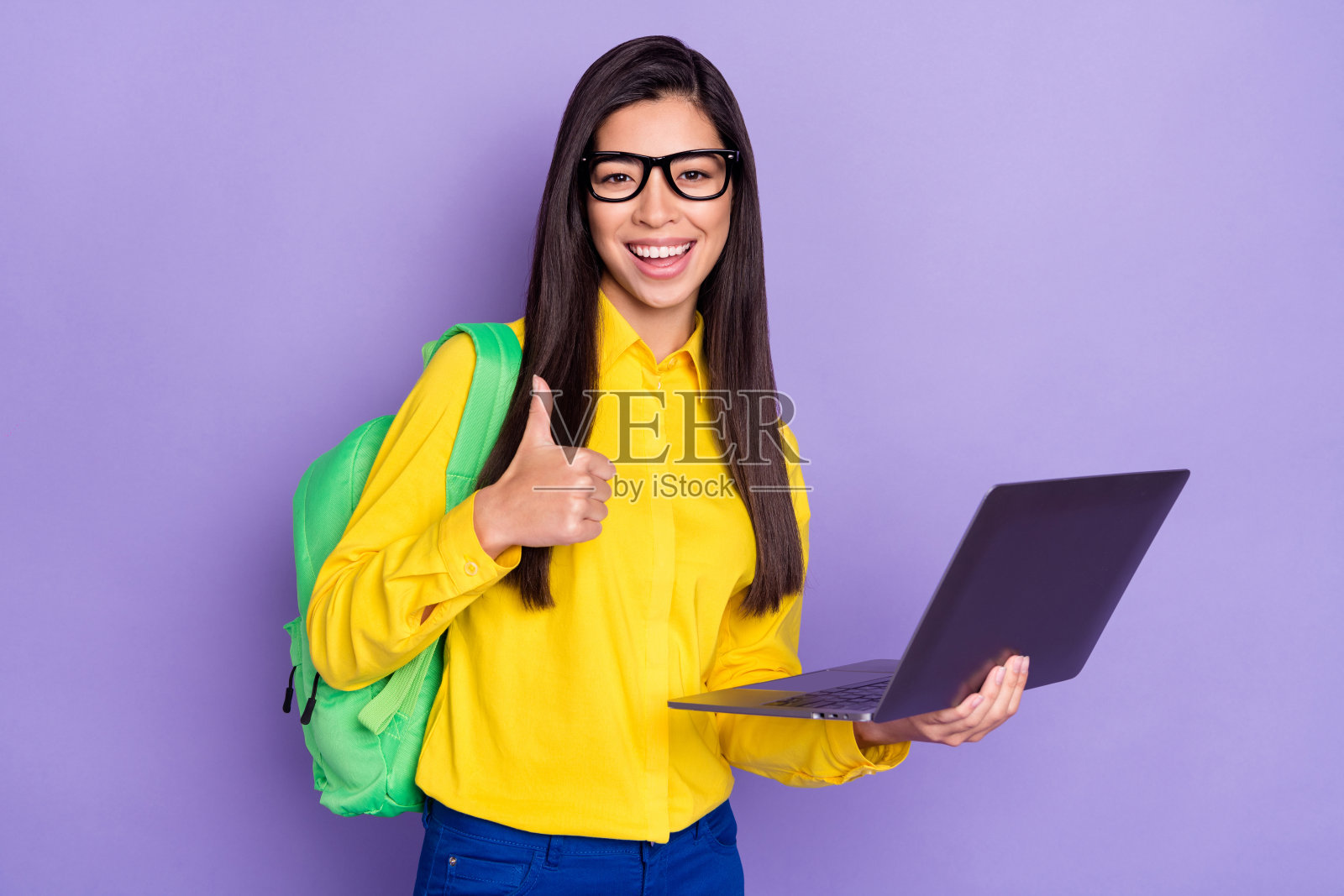 身穿黄色衬衫、竖起大拇指的迷人年轻棕发女子,手持笔记本电脑,背景为紫色。照片摄影图片