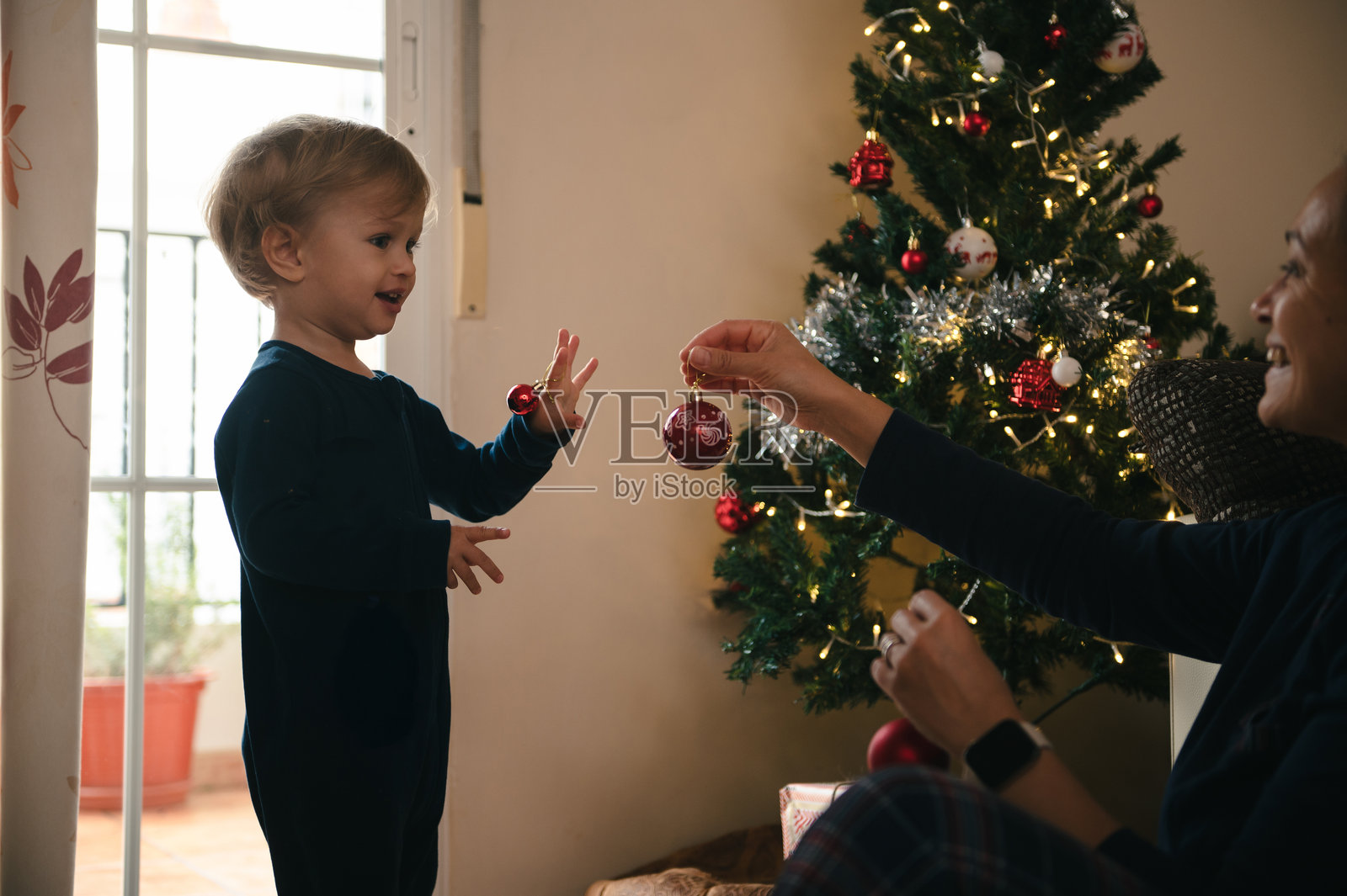 温馨的家中,孩子开心地伸手去够挂在闪烁着灯光的圣诞树上的装饰品照片摄影图片