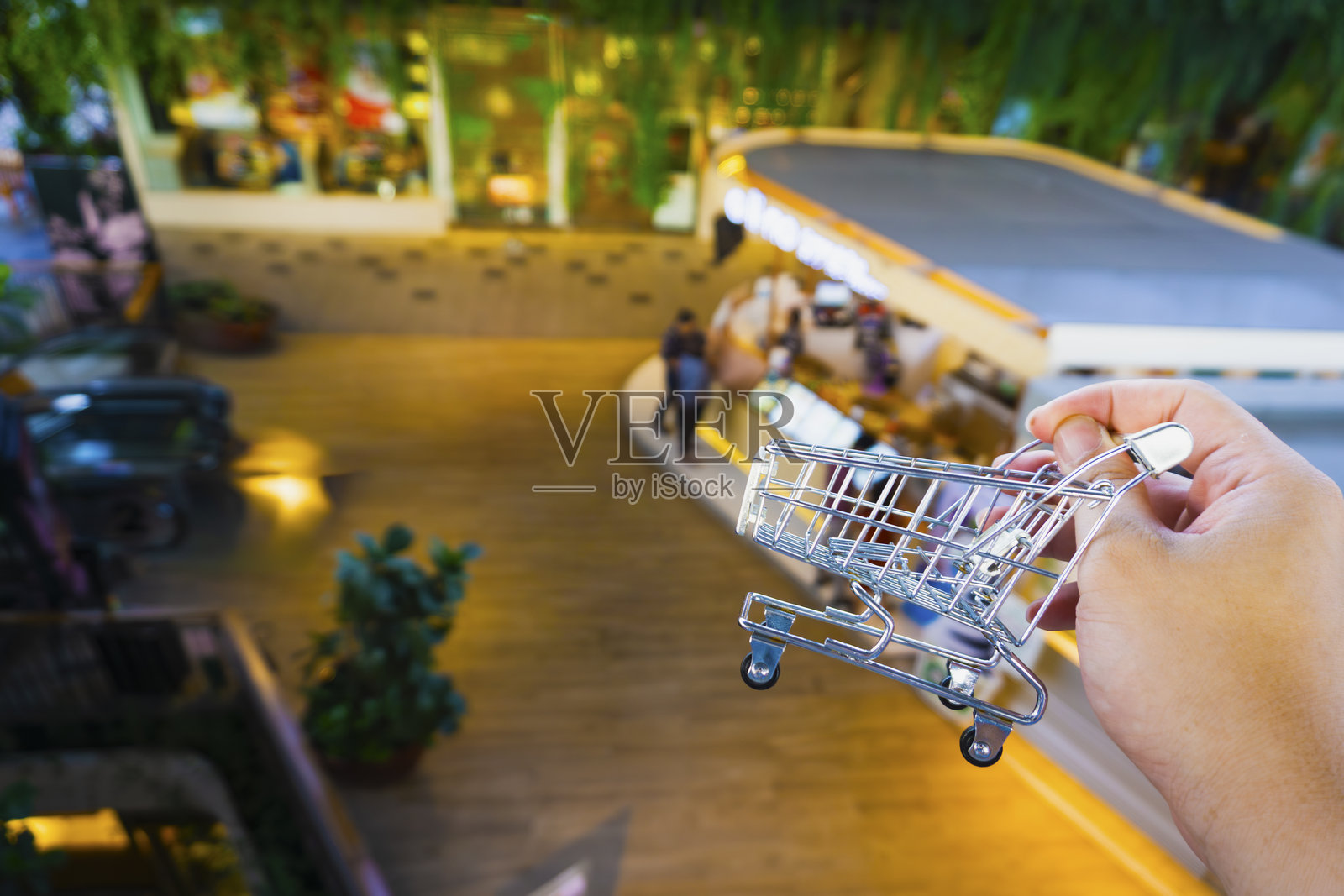 手持微型金属购物篮模型,背景为模糊的现代购物中心,象征着消费主义、在线购物、电子商务和零售营销策略。照片摄影图片