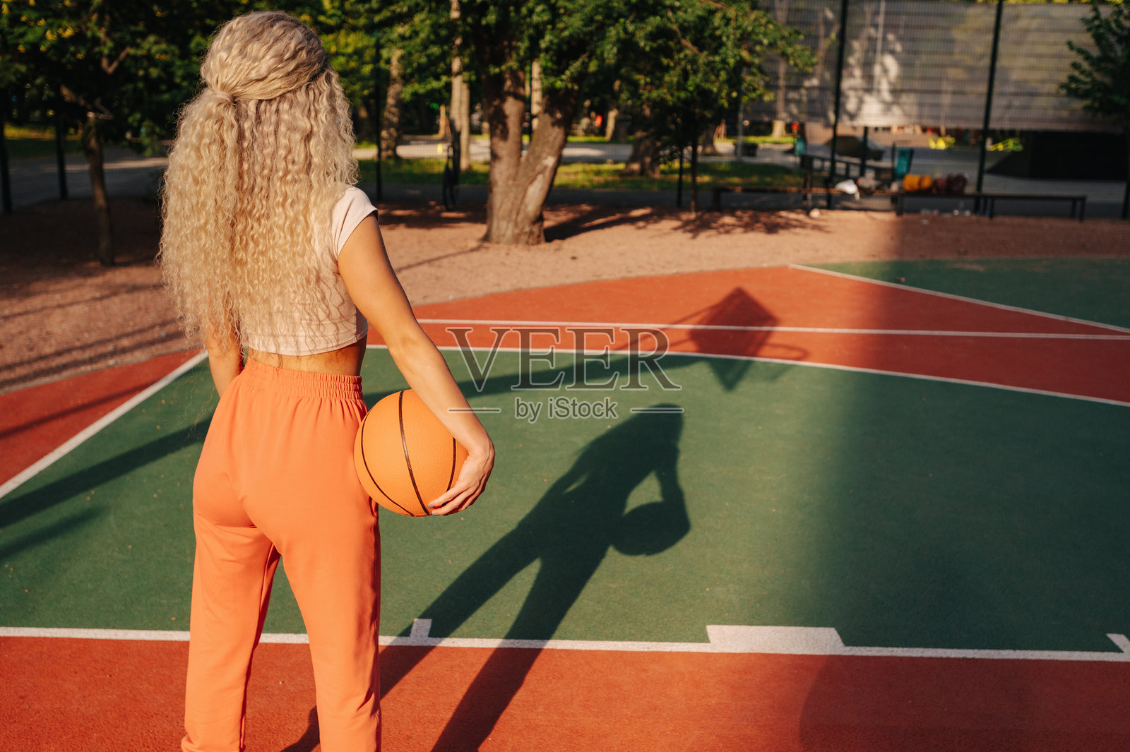 身穿运动服的女性在户外篮球场上的背影,明亮的阳光在她身后投下长长的身影,地面色彩鲜艳。照片摄影图片