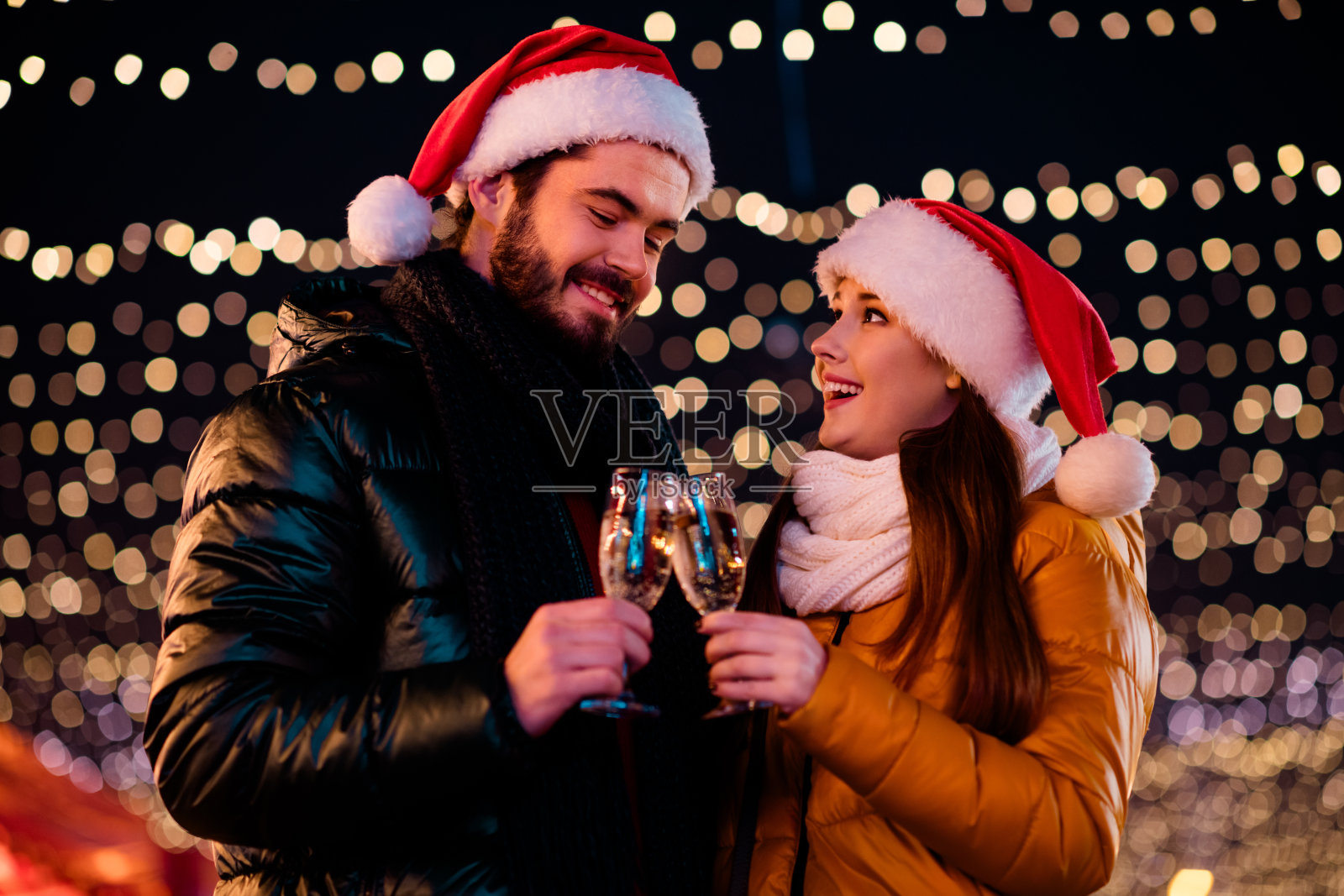 在闪烁的灯光下,一对情侣在平安夜举杯庆祝,他们穿着温暖的外套,享受着彼此的爱意和节日的欢乐。照片摄影图片