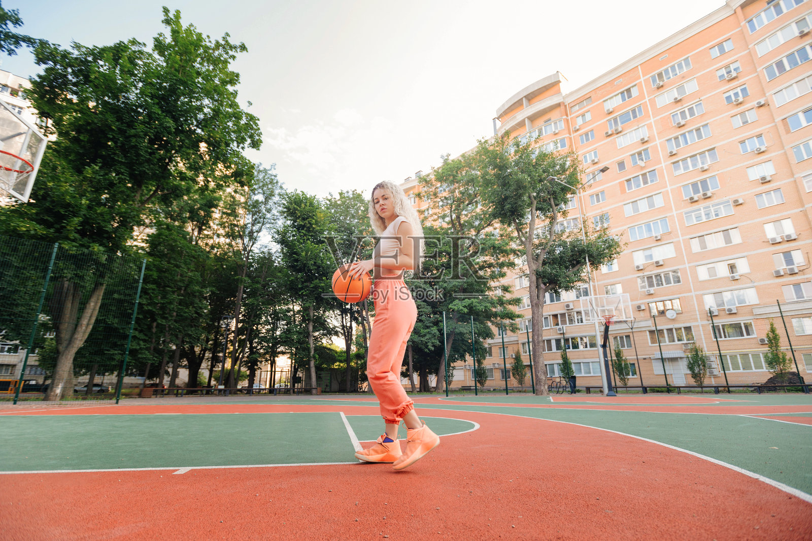 年轻女子身穿珊瑚色裤子和露脐上衣,手持篮球,在夏日阳光下的户外球场上,自信满满,准备比赛。照片摄影图片