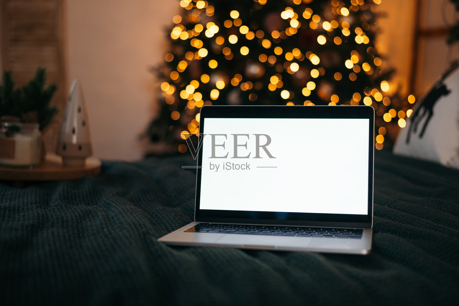 舒适的床上放着一台屏幕空白的笔记本电脑,背景是亮着温暖灯光的圣诞树,营造出节日的家庭氛围。照片摄影图片