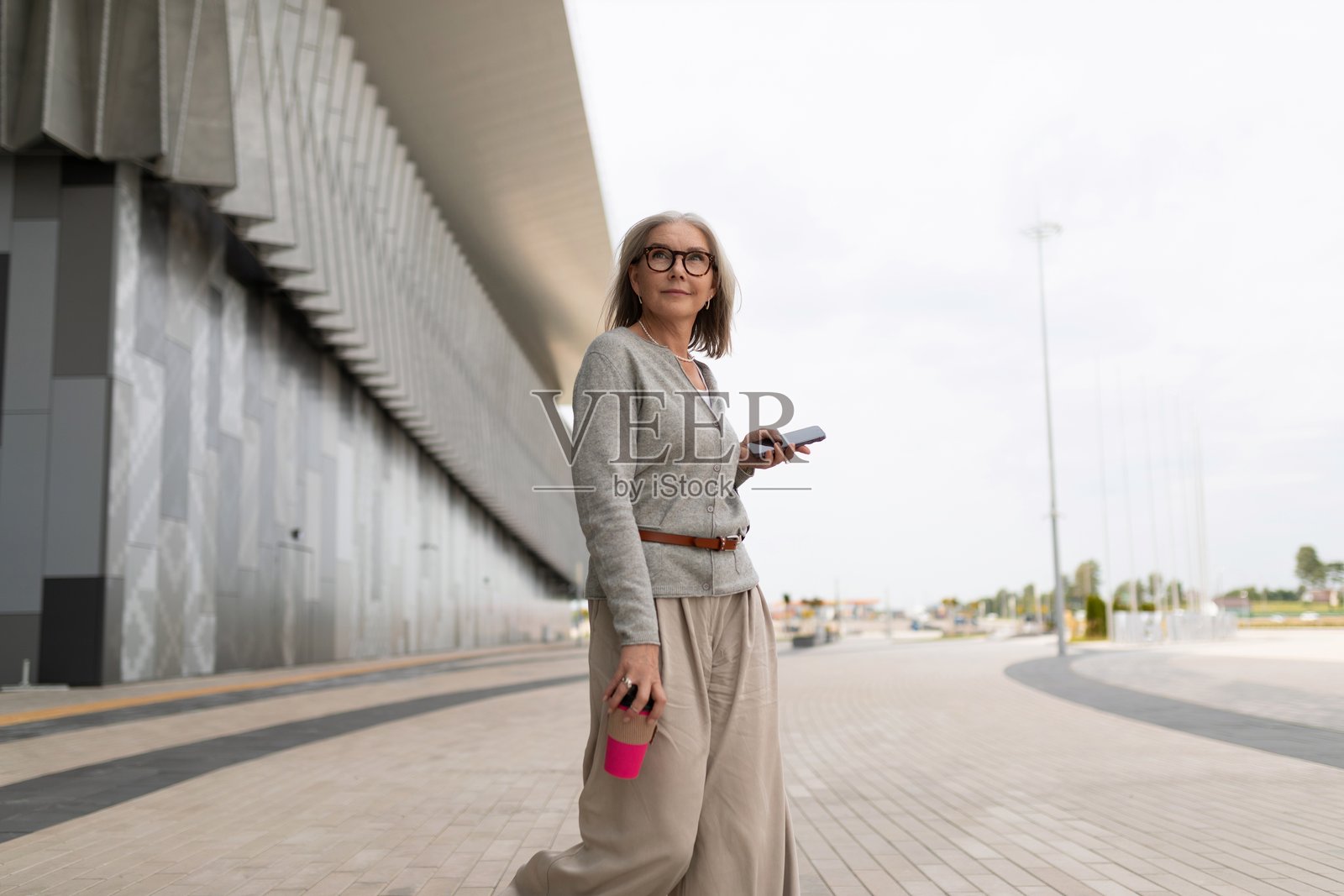 一位成熟的女性在现代都市建筑旁,手持咖啡杯和智能手机散步。照片摄影图片