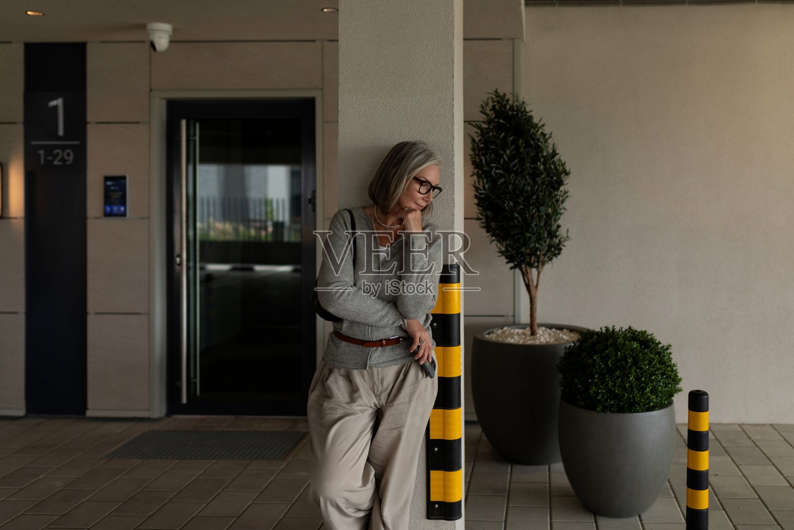 一位年长的女性白天靠着柱子,在现代建筑外等候。照片摄影图片