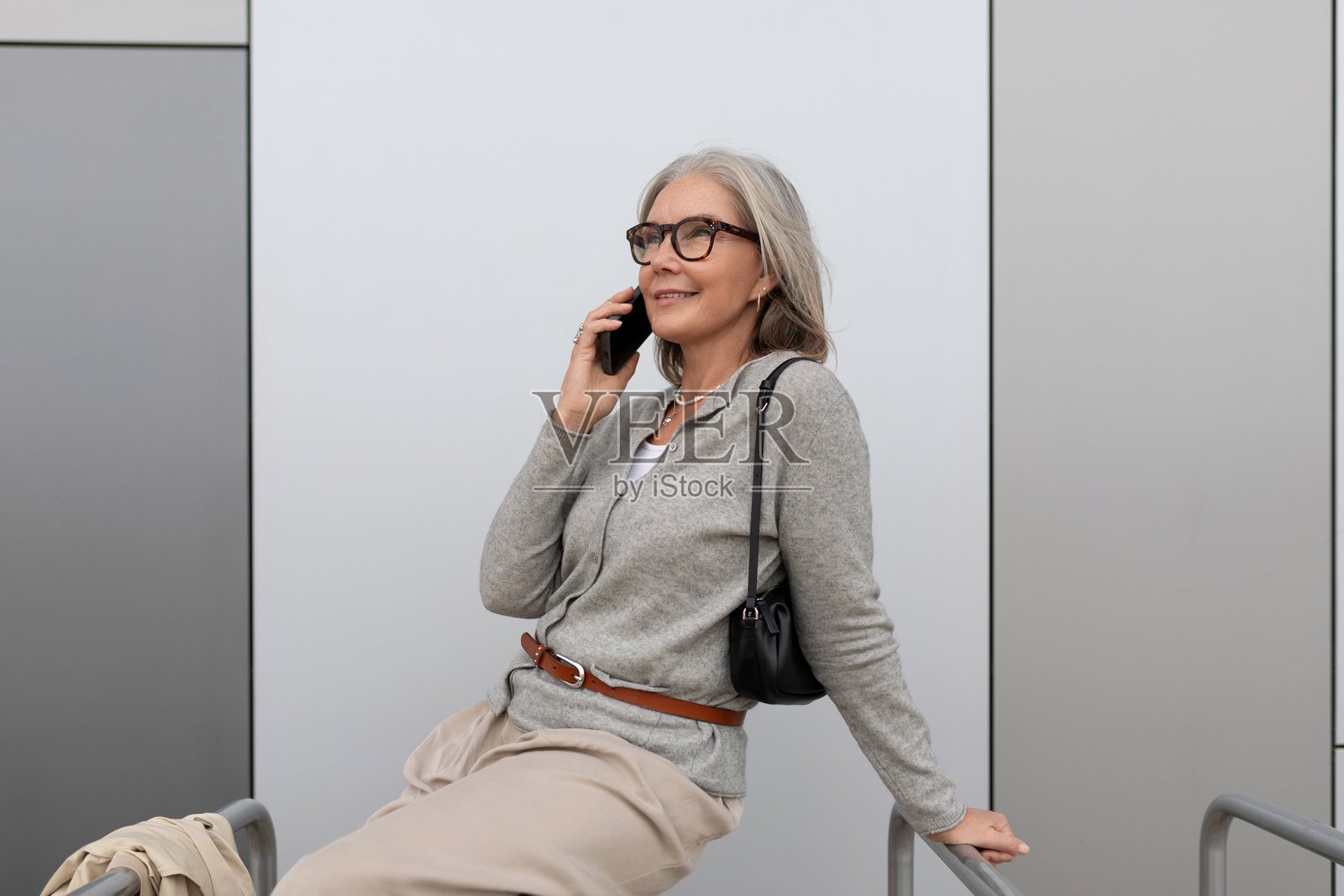 身着休闲装的老年女性,在都市背景下,倚靠着现代墙壁,正在打电话。照片摄影图片