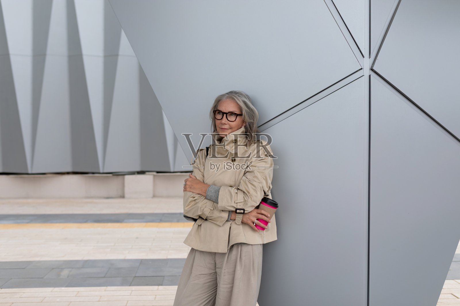 一位时尚的年长女性倚靠在现代建筑旁,在都市环境中享用咖啡。照片摄影图片