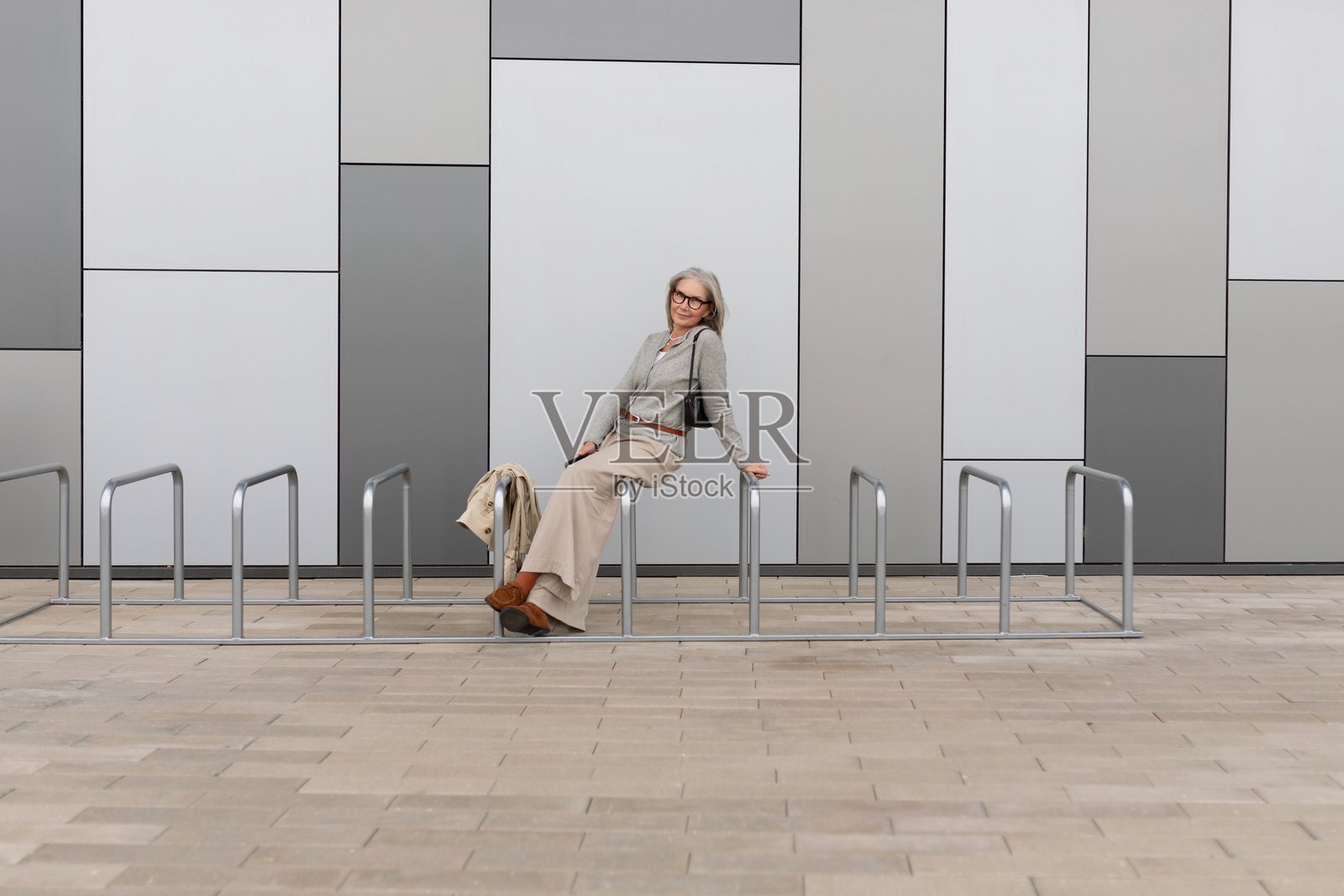 时尚的年长女性在白天的现代都市环境中,倚靠在自行车架上放松。照片摄影图片