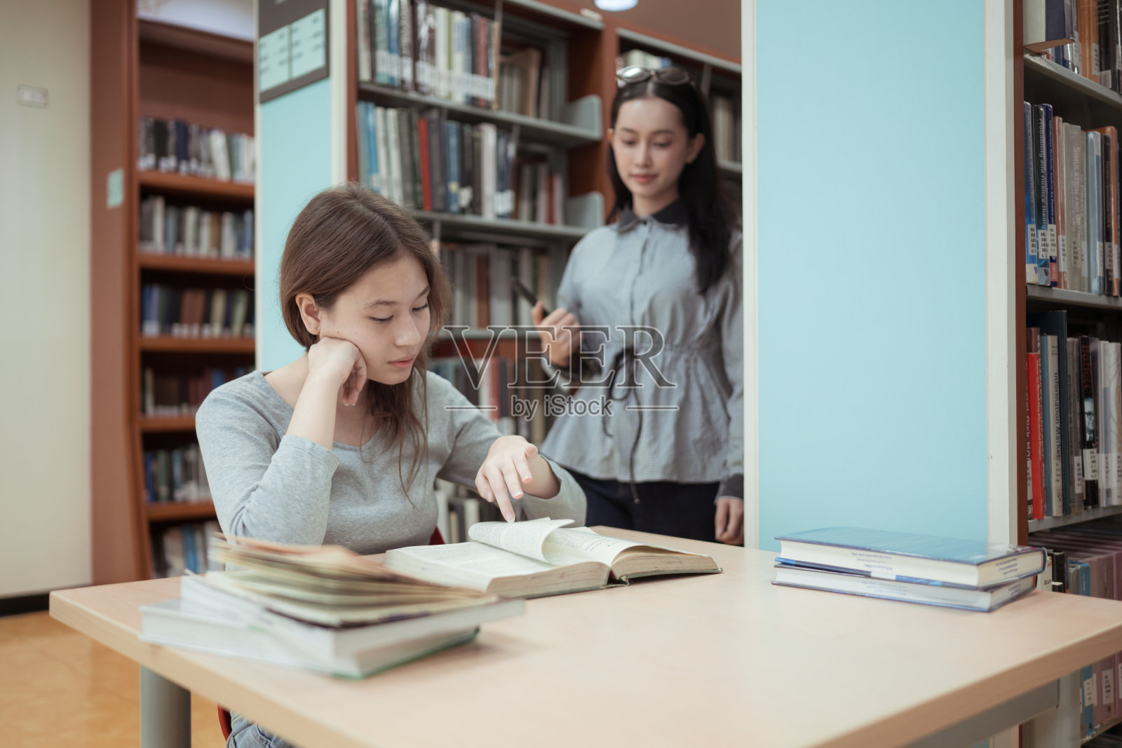两位年轻女士在图书馆里,手持书籍和电子平板,开心地微笑着交谈。照片摄影图片