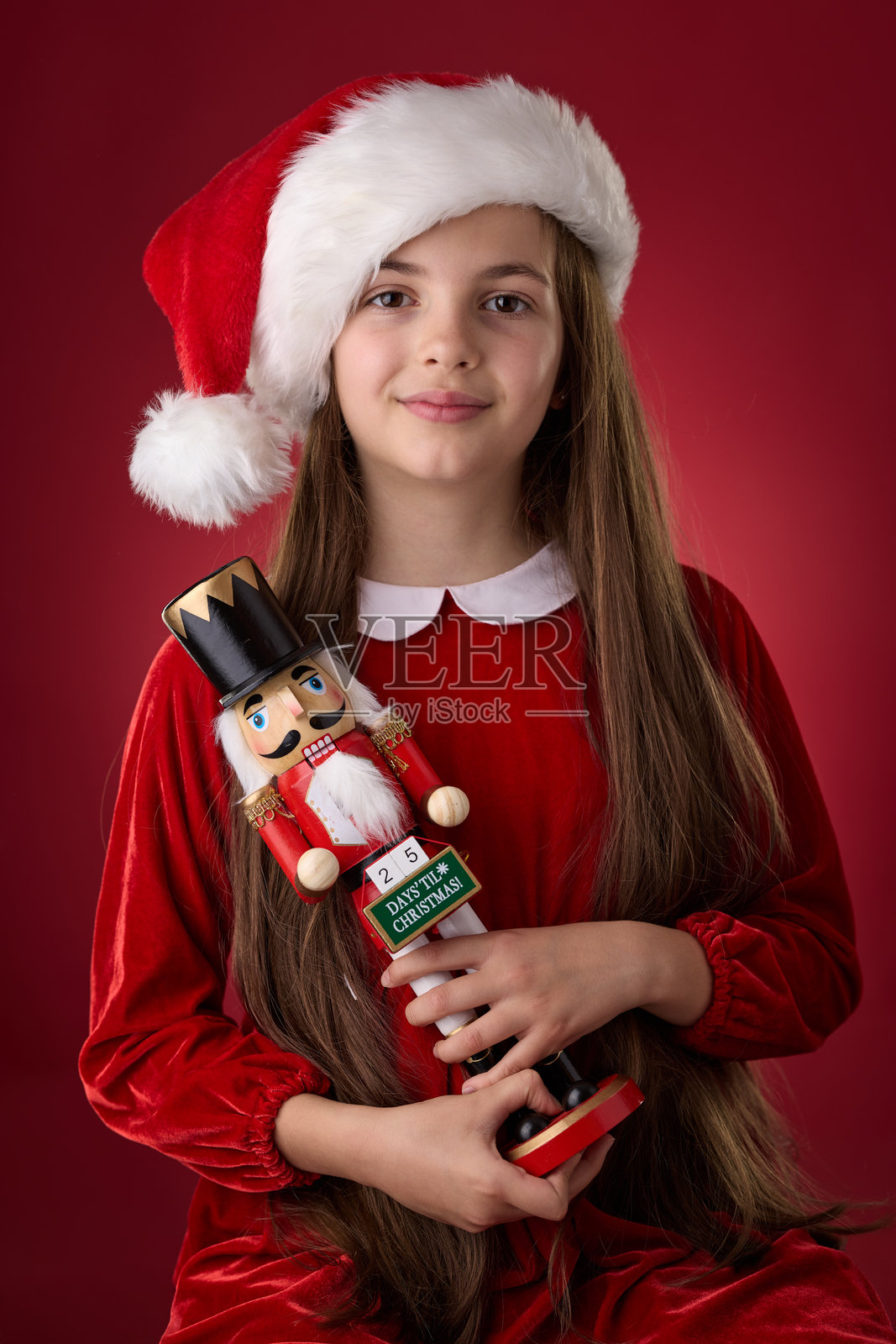 身穿红色圣诞帽的小女孩肖像,手持胡桃夹子木制圣诞玩具,背景为红色照片摄影图片