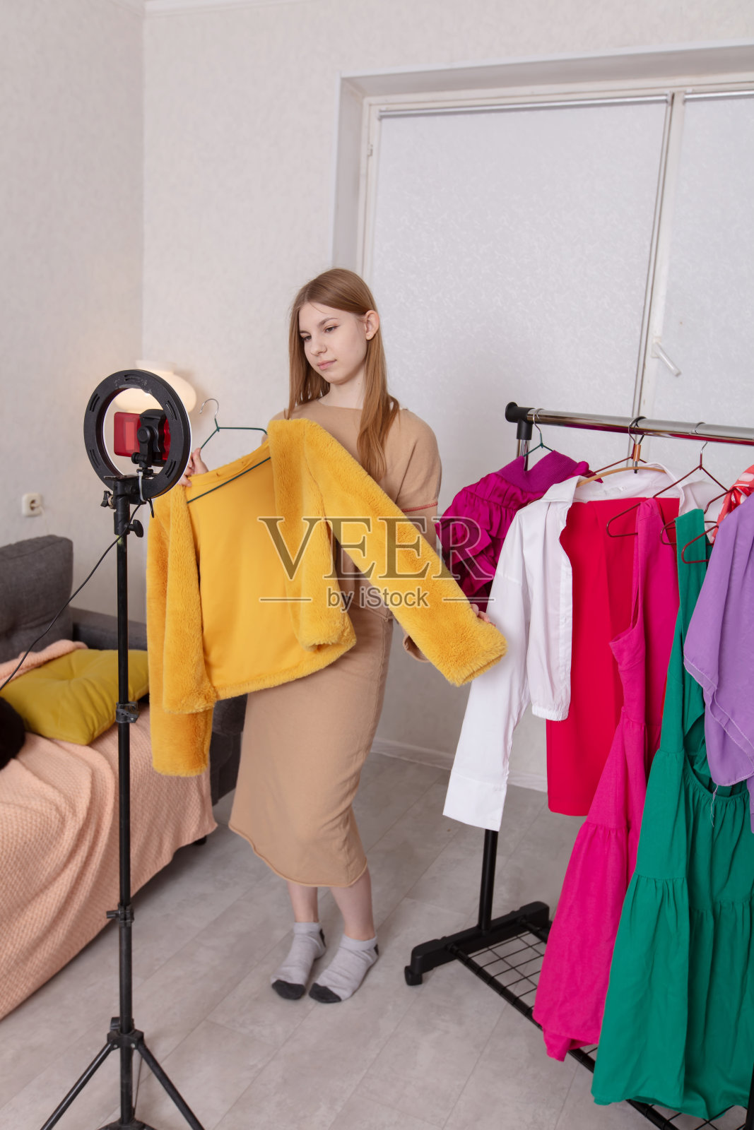 年轻女子在家中明亮的房间里,在挂满彩色服装的衣架旁,搭配服装进行拍摄。照片摄影图片