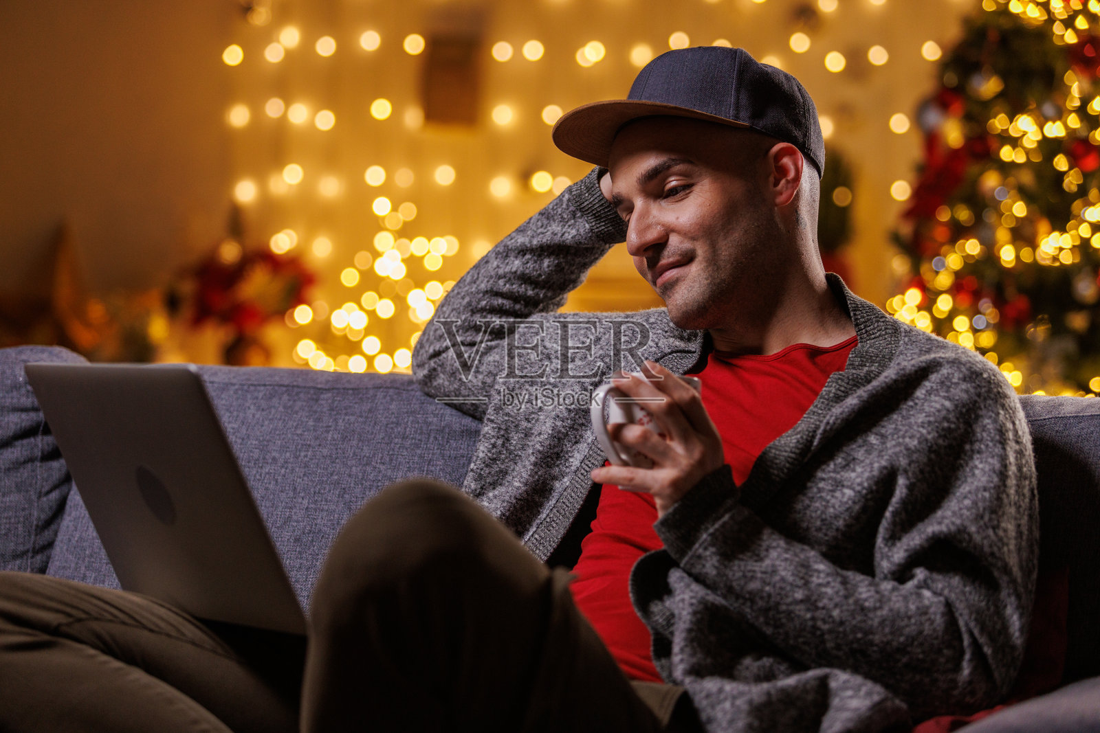 舒适的男人在圣诞灯饰下,在家中的沙发上使用笔记本电脑,手捧马克杯照片摄影图片