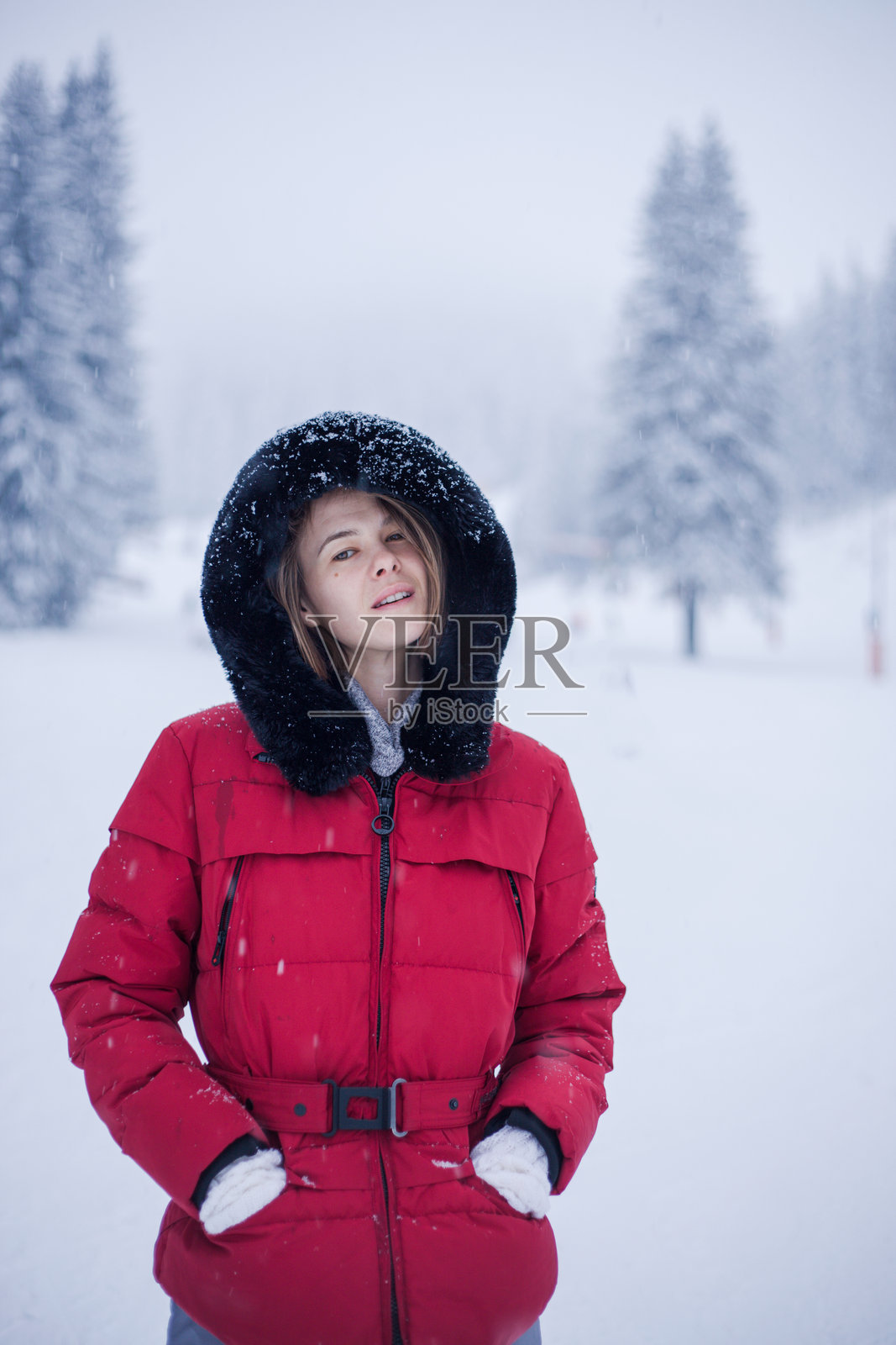 身穿红色冬季连帽夹克的自然美女的肖像,她站在雪地景观中,周围飘着雪花。照片摄影图片