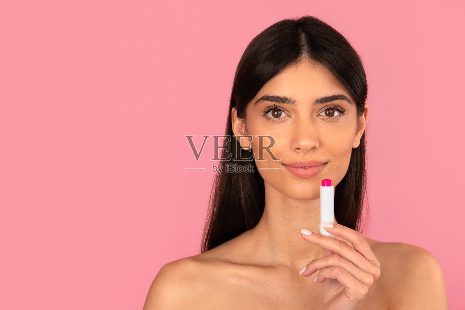 一位美丽的年轻女子,在粉色背景前,嘴唇微微上扬,正在涂抹润唇膏。照片摄影图片