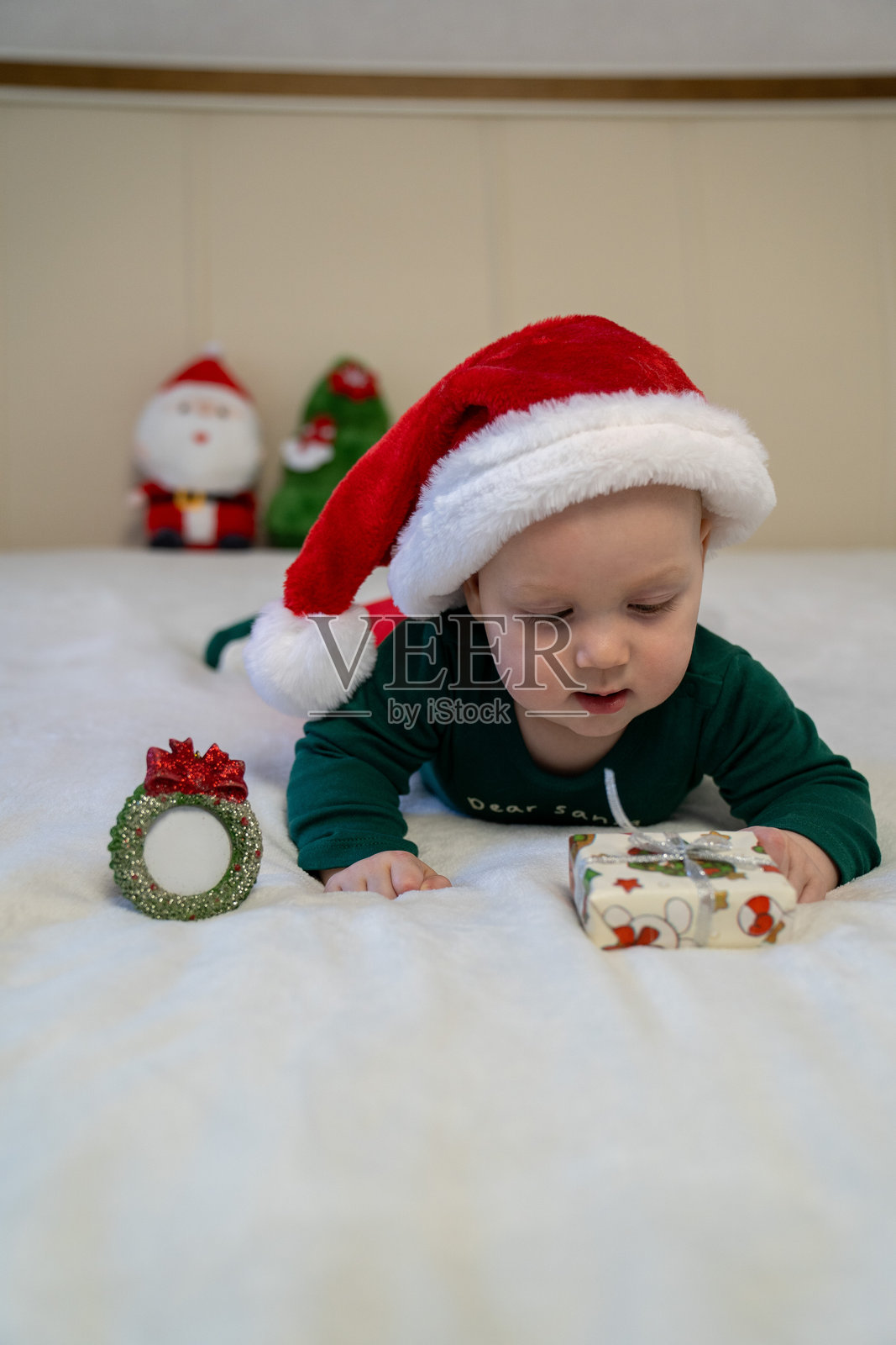 穿着圣诞老人帽子和绿色节日服装的婴儿躺在白色床上,伸手去拿一个小包装好的圣诞礼物。婴儿蹒跚学步,庆祝第一个圣诞节和新年夜。照片摄影图片