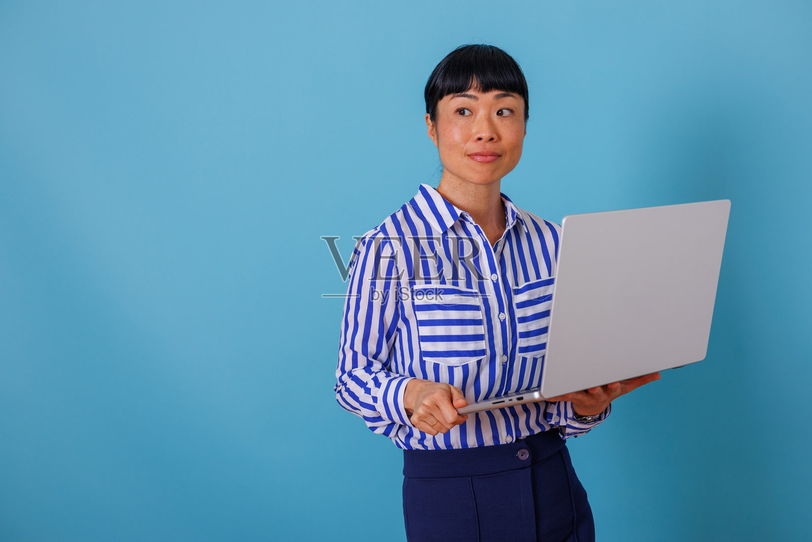 亚洲女商人手持笔记本电脑,站在蓝色背景前,望向远方,思考着商业成功与创新。照片摄影图片