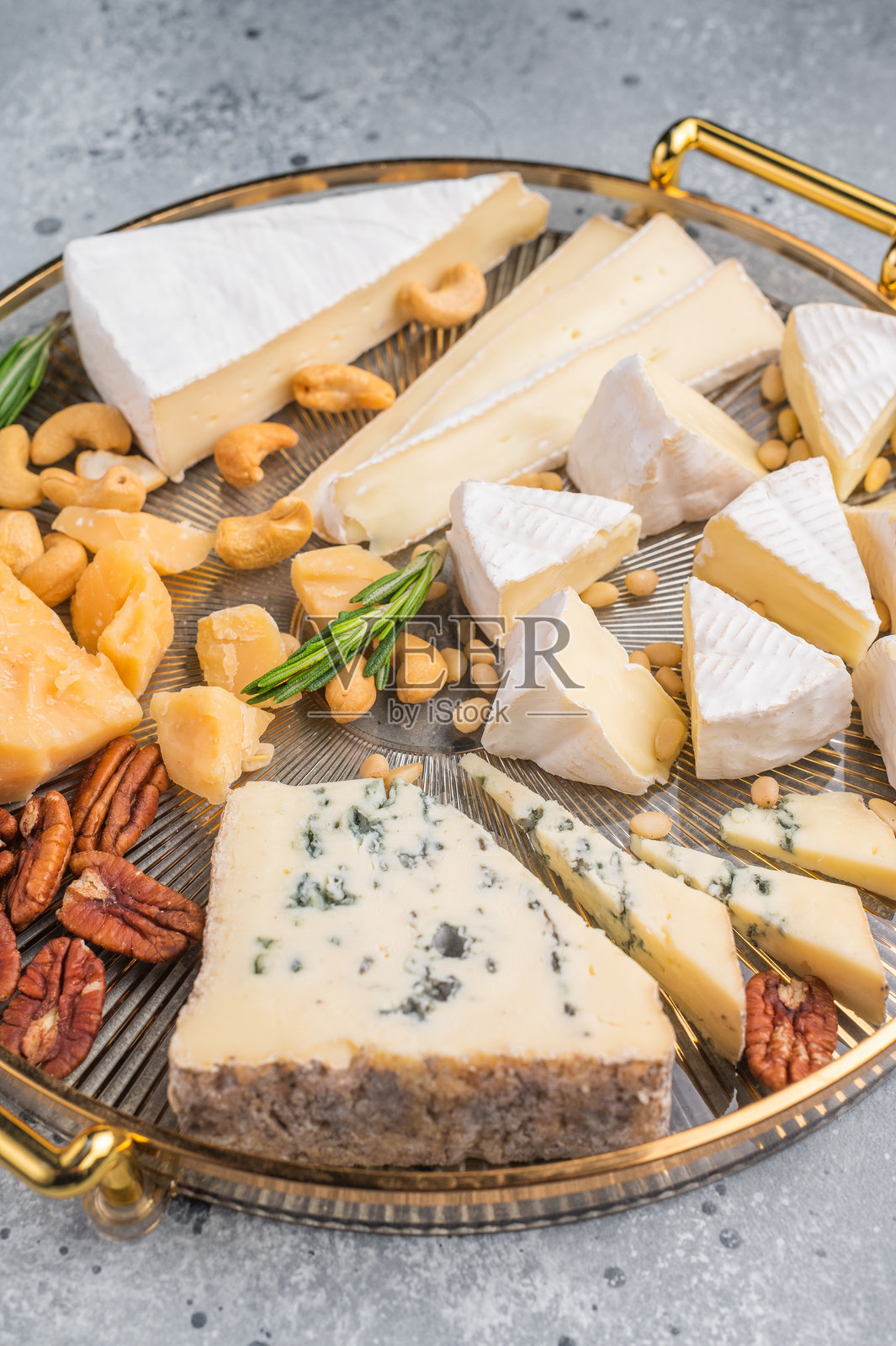 金色托盘上的精致奶酪拼盘,配有坚果和香草,置于现代灰色混凝土背景前,尽显奢华美食风范。照片摄影图片