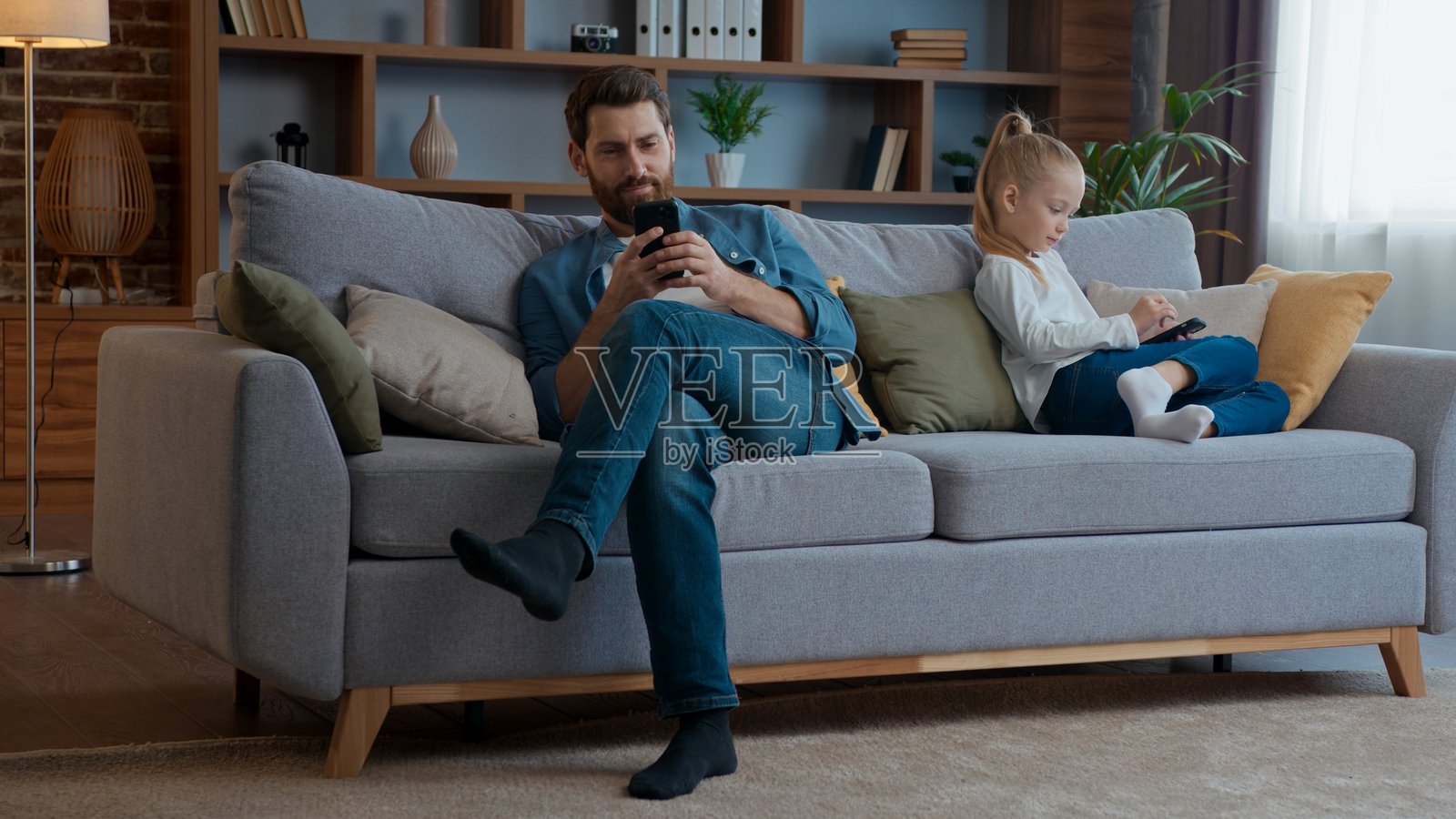 白种人家庭的电子产品成瘾问题,一个小女孩和爸爸在家里的客厅沙发上,爸爸正在滑动手机应用,孩子和爸爸各玩各的电子设备,孩子是女儿,爸爸是daddy照片摄影图片