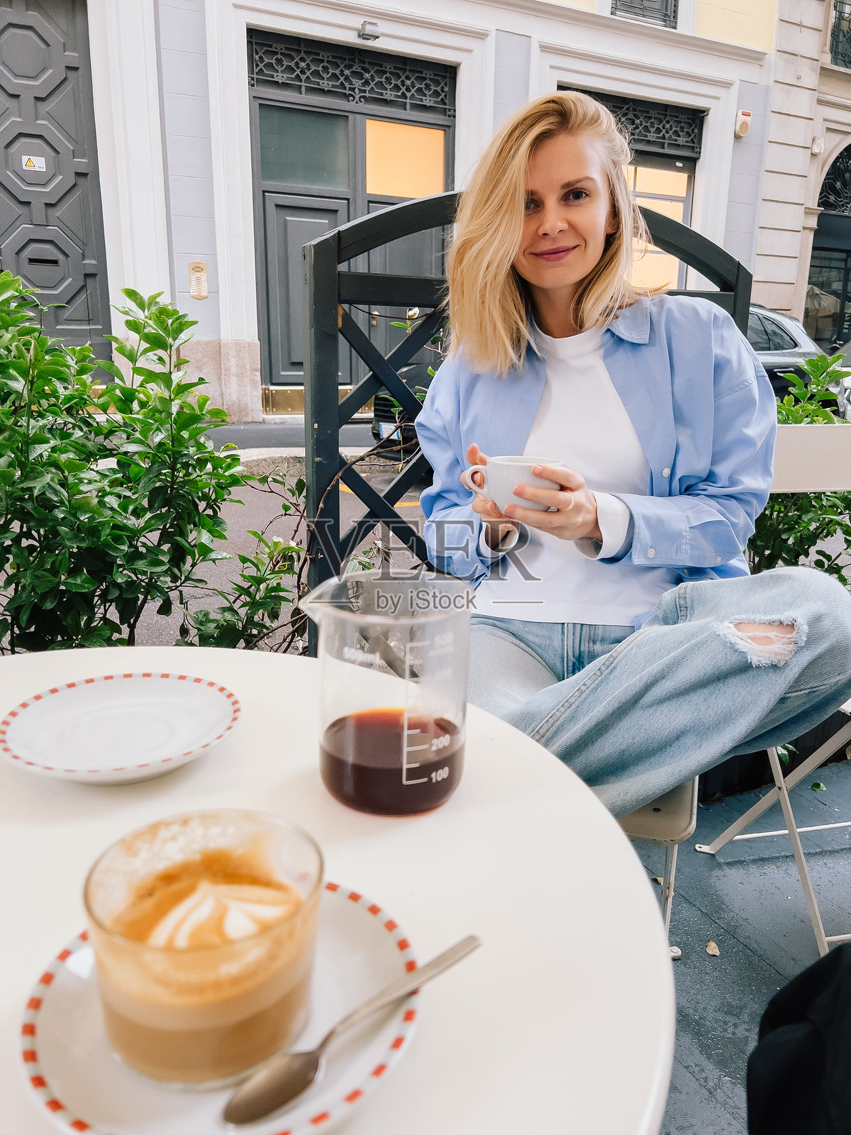 年轻女子坐在熙熙攘攘的街边咖啡馆的桌旁,身着休闲装,品尝着浓郁的咖啡和奶泡丰富的卡布奇诺,享受着充满活力的都市氛围。女子正在啜饮卡布奇诺。照片摄影图片
