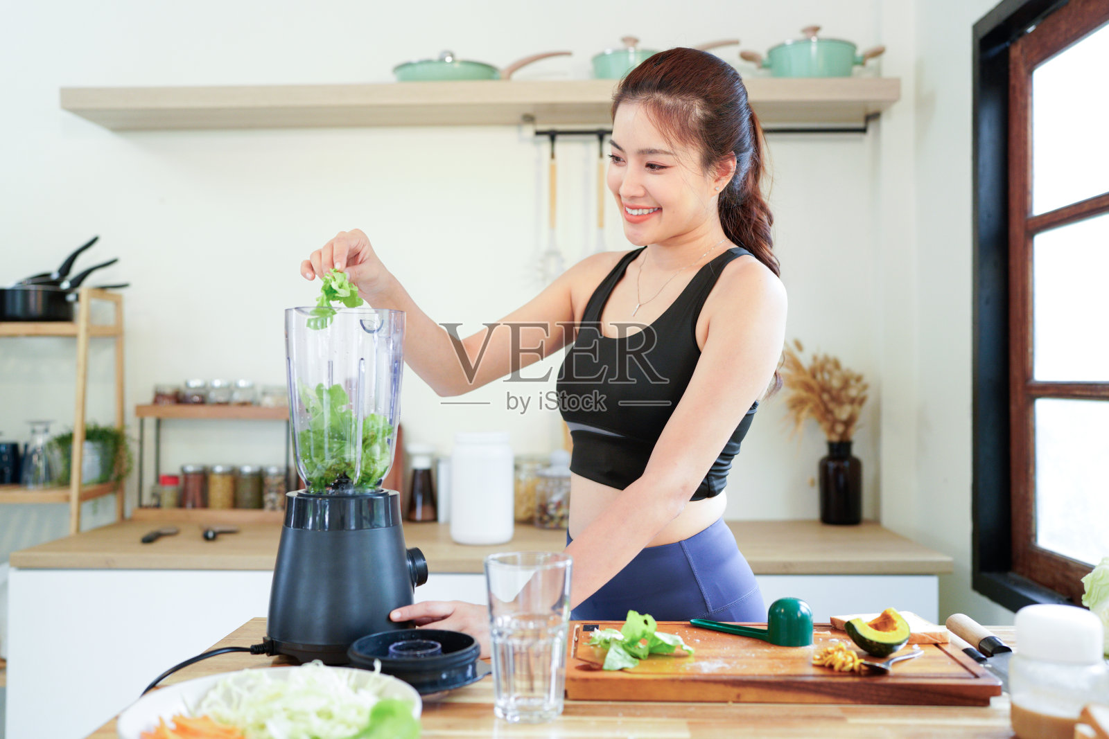 健康食品。身穿休闲运动服的女性在家中厨房用搅拌机制作健康冰沙,在制作绿色冰沙时将新鲜沙拉叶放入搅拌机照片摄影图片