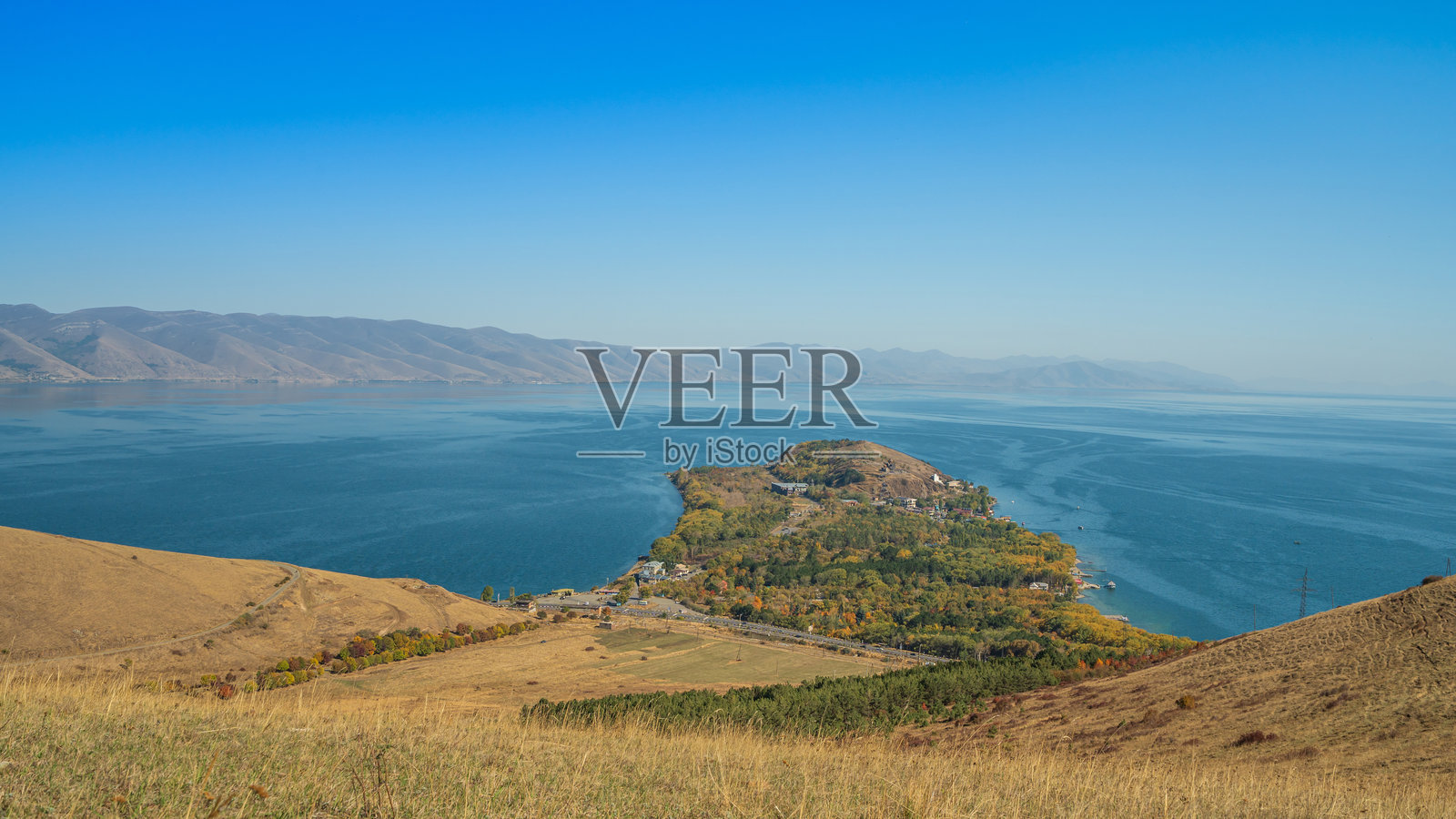 亚美尼亚塞凡湖的风景,湖上有个小半岛,周围是清澈的蓝色湖水和金黄色的干枯山丘,天空湛蓝明亮。照片摄影图片