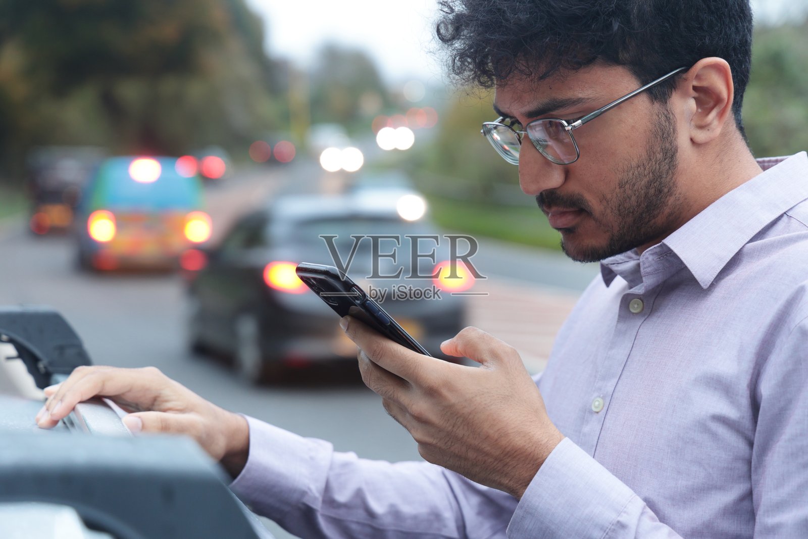 年轻的商人坐在公路旁损坏的汽车旁,正在使用手机照片摄影图片