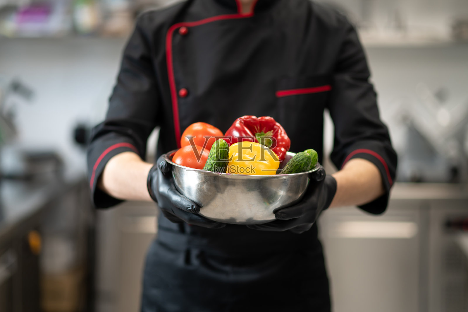 一位穿着厨师服、身份不明的人站着,手中拿着一道包含胡椒、黄瓜和番茄的菜肴,背景是餐厅厨房照片摄影图片