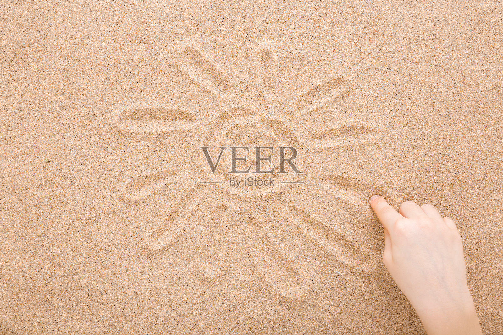 特写镜头,孩子的手指在干燥的浅棕色海沙上画太阳和光芒。夏季概念。俯视角度。照片摄影图片