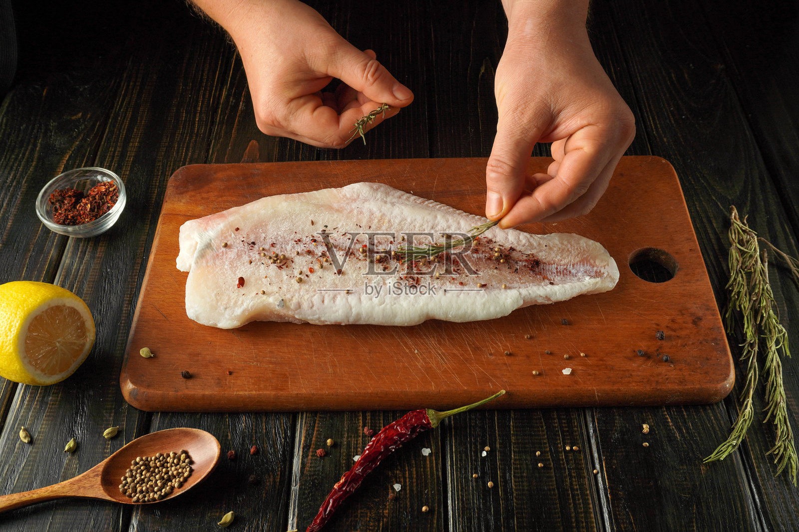 厨师正在木质砧板上精心为新鲜鱼片撒上香料和香草。柠檬和各种香草放在旁边,突出了烹饪的活动。照片摄影图片
