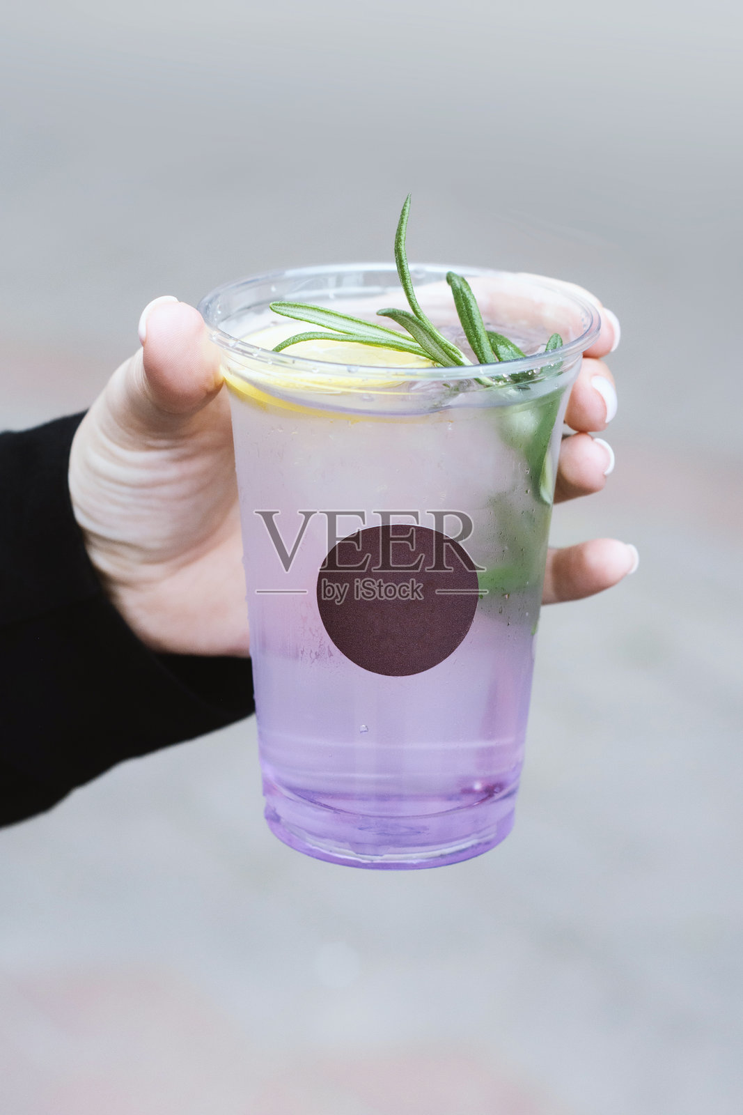 女性手持一杯带有新鲜迷迭香装饰的紫色渐变鸡尾酒或柠檬水,盛放在透明玻璃杯中。照片摄影图片