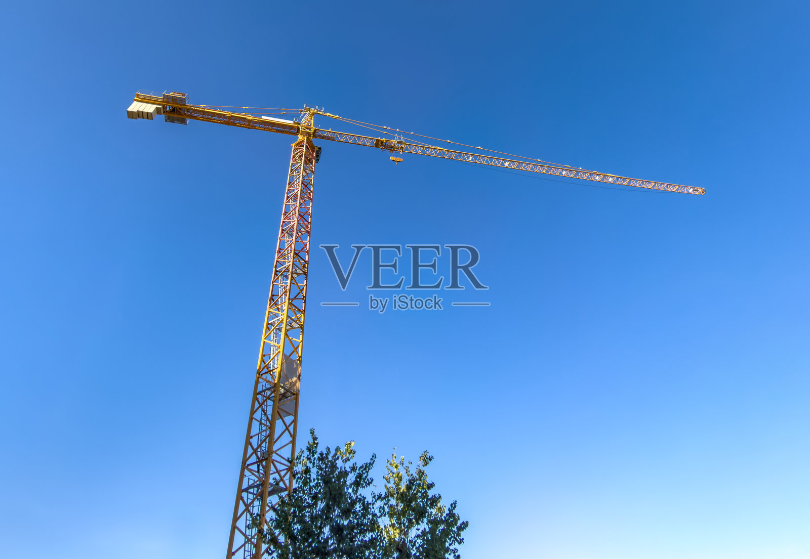 在明亮的日光下,从下方仰视,高耸的黄色塔式起重机架设在晴朗的蓝天上,背景中可见树梢。照片摄影图片