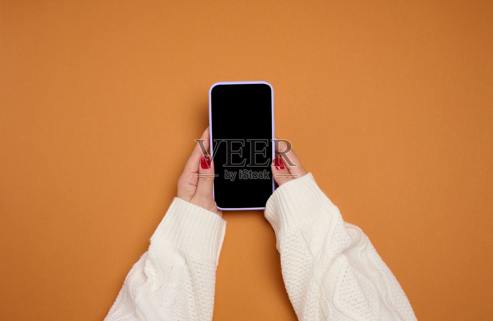 身穿白色针织毛衣的女性双手持有一部黑色屏幕的手机,背景为橙色和棕色。照片摄影图片