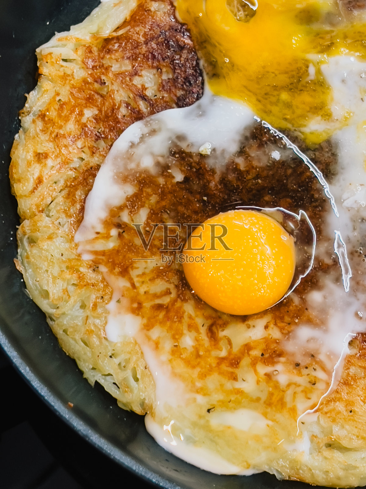 特写:煎锅中正在烹制的酥脆土豆煎饼,上面放着一个煎蛋,制作出诱人的美味早餐。铁锅里的酥脆土豆煎饼配太阳蛋。照片摄影图片