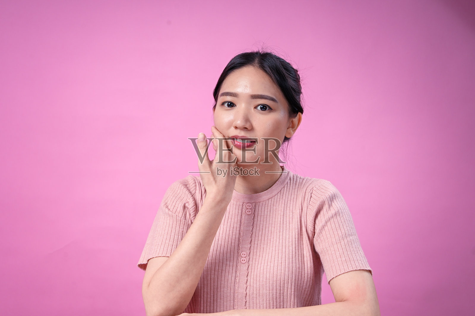 一位黑发盘成整洁发髻的年轻亚洲女性,身穿罗纹粉色短袖上衣,面带灿烂笑容,手托下巴,在粉色纯色背景前摆出开朗的姿势。照片摄影图片