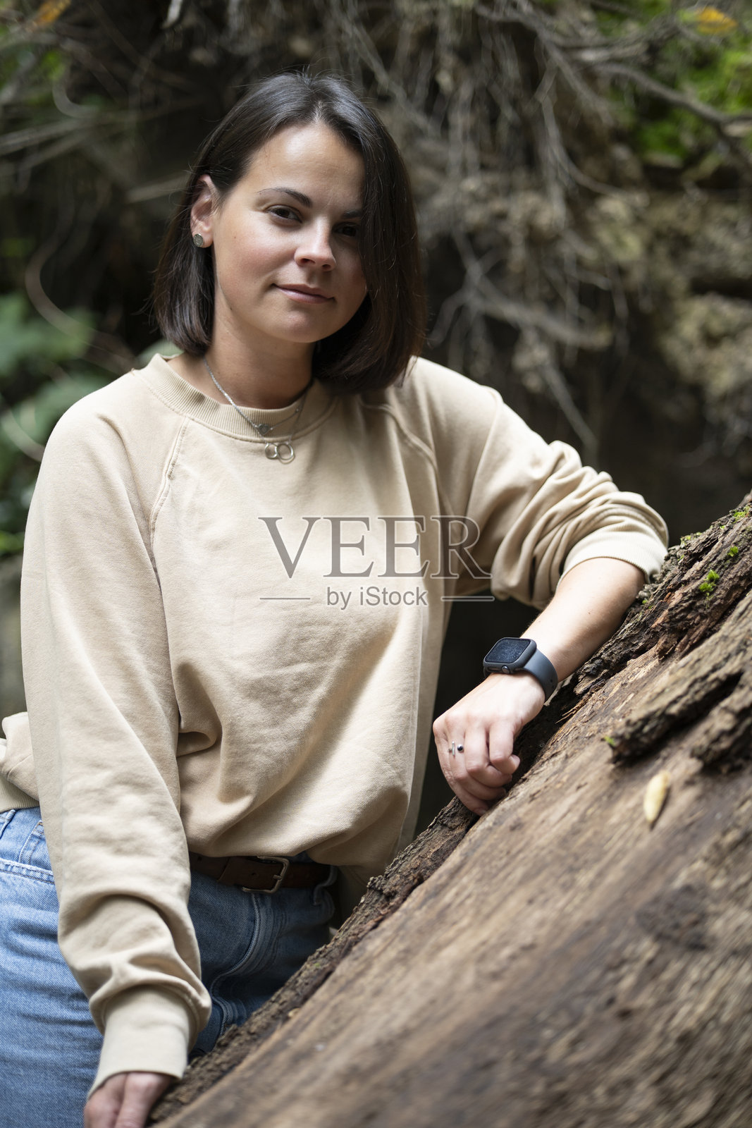 身穿卫衣的微笑女孩的垂直肖像,倚靠在茂密的森林树干旁,柔和的自然光,模糊的背景,可供复制的空间。和谐、温暖、与自然连接的概念。照片摄影图片