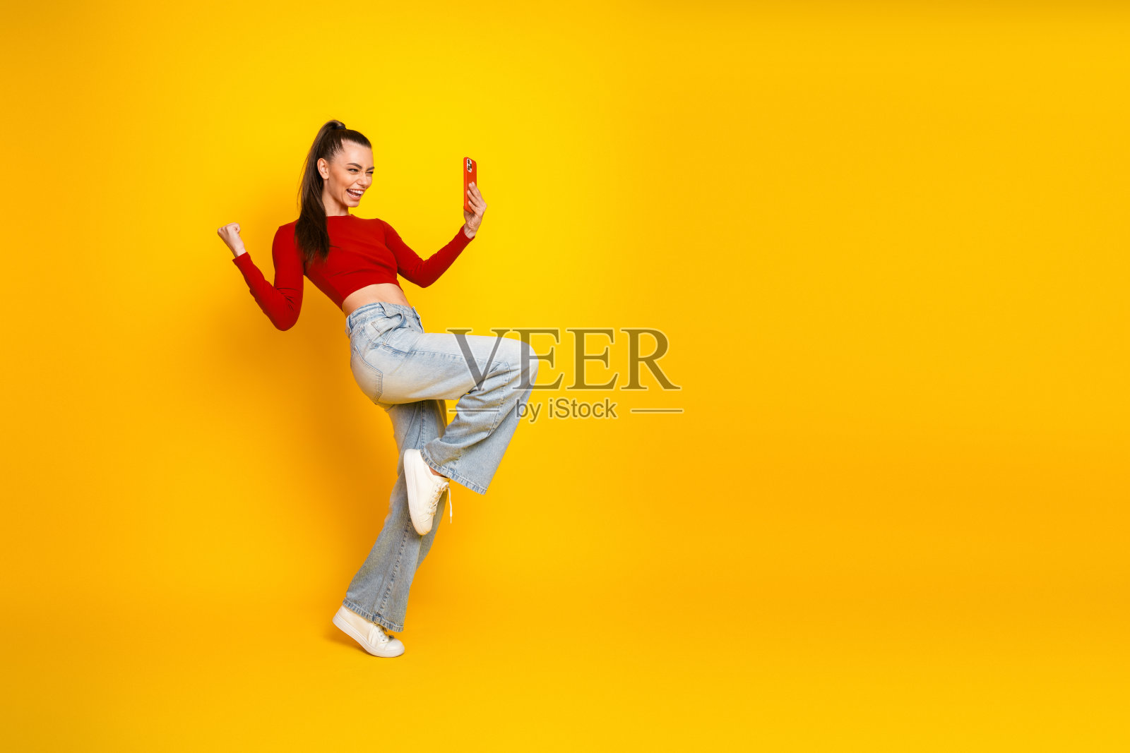 一位穿着红色露脐上衣和牛仔裤的年轻女子,背景是明亮的黄色,她微笑着自拍,适用于时尚生活方式广告和购物概念照片摄影图片