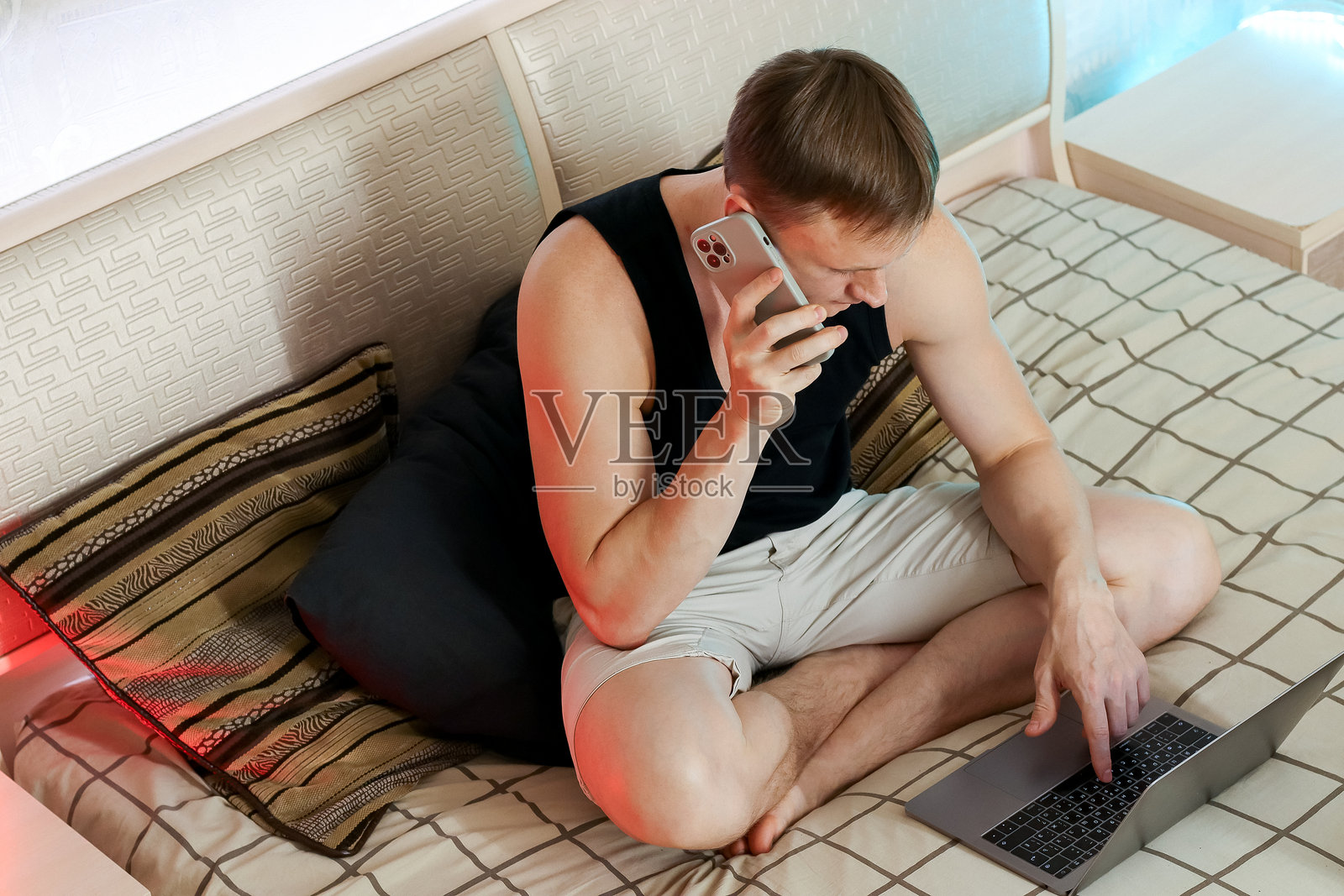 年轻男子在床上同时处理多项事务,包括打电话和用笔记本电脑打字照片摄影图片