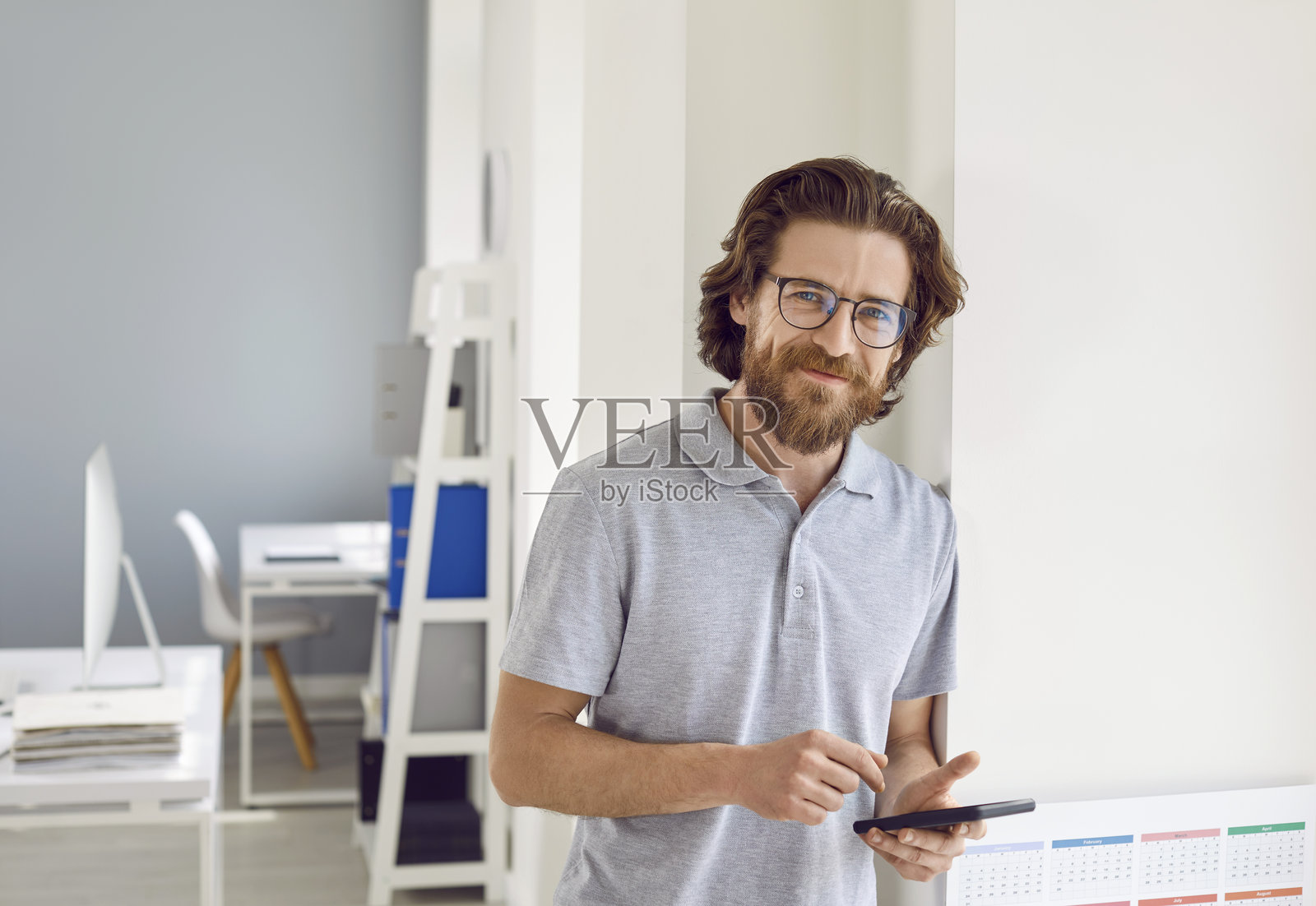 穿着T恤的快乐商务人士站在办公室里,手里拿着手机,面带微笑照片摄影图片