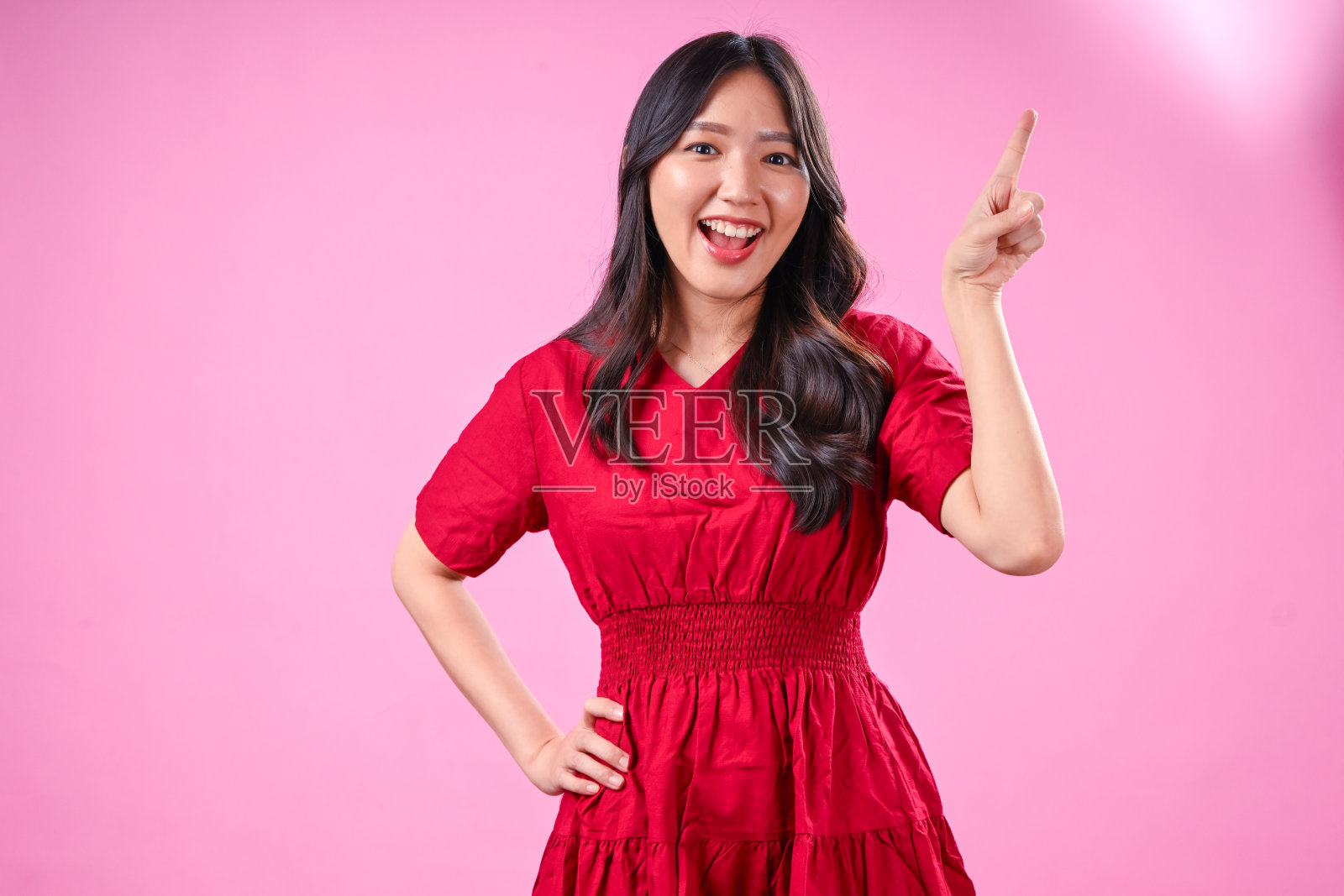 一位拥有黑色波浪长发的亚洲年轻美女,身穿红色连衣裙,面带微笑,一只手自信地向上指着,背景为粉色,暗示着一个想法。照片摄影图片