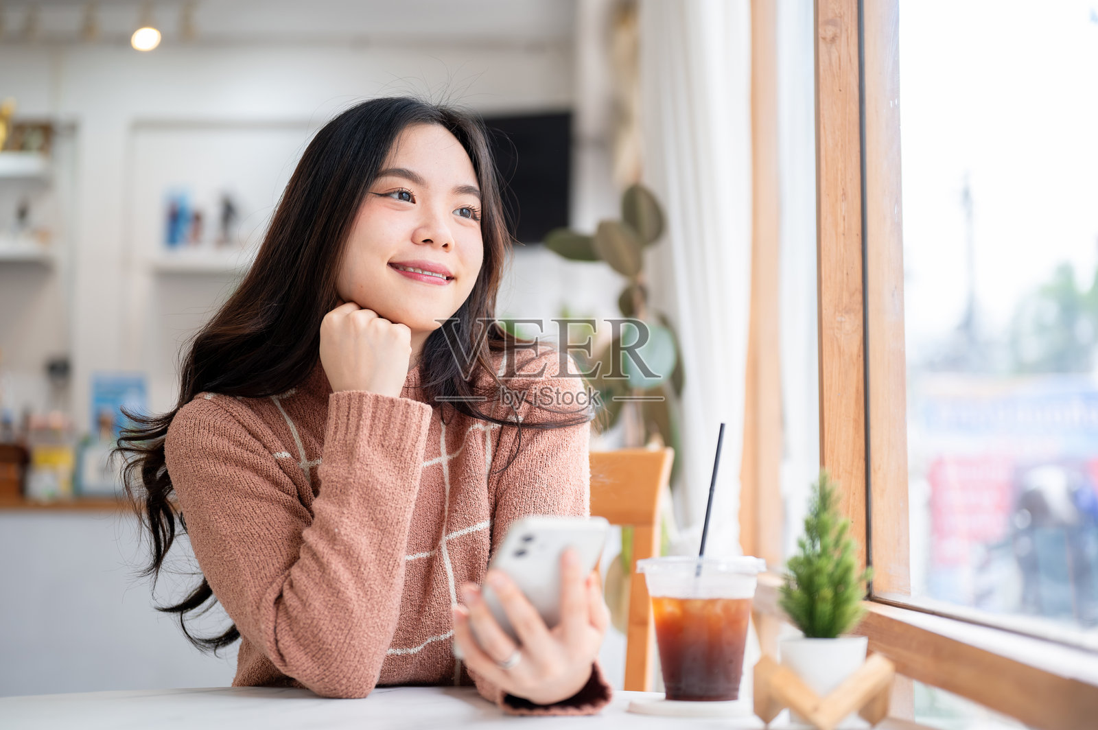 亚洲女性坐在咖啡馆或咖啡店的玻璃墙旁桌边,手持手机,目光望向别处。照片摄影图片