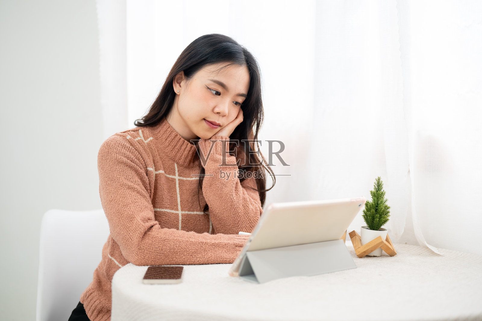 亚洲女性在白色房间或咖啡馆的桌旁,手托着头,手持钢笔在平板电脑上书写。照片摄影图片